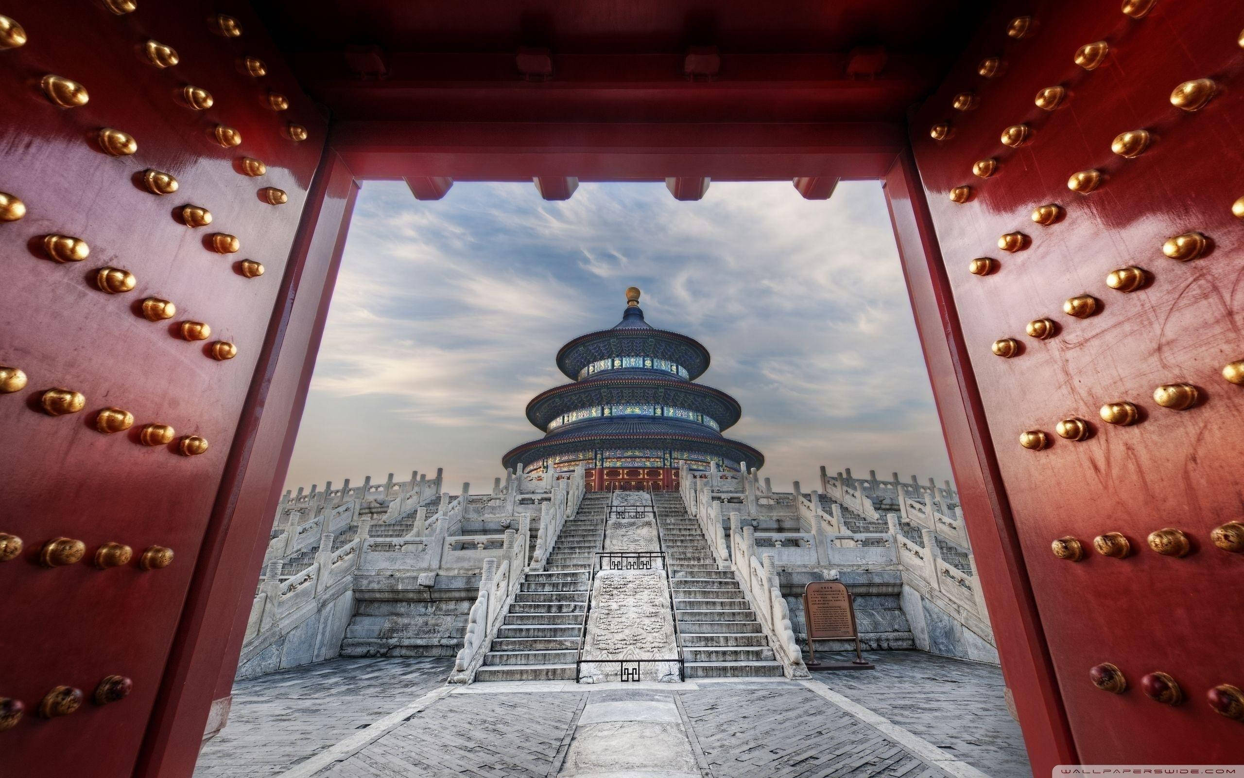 Beijing 桌布 壁紙 — 一座宏偉的傳統中式寺廟，以帶有金色門釘的紅色木門框住，天空部分多雲。