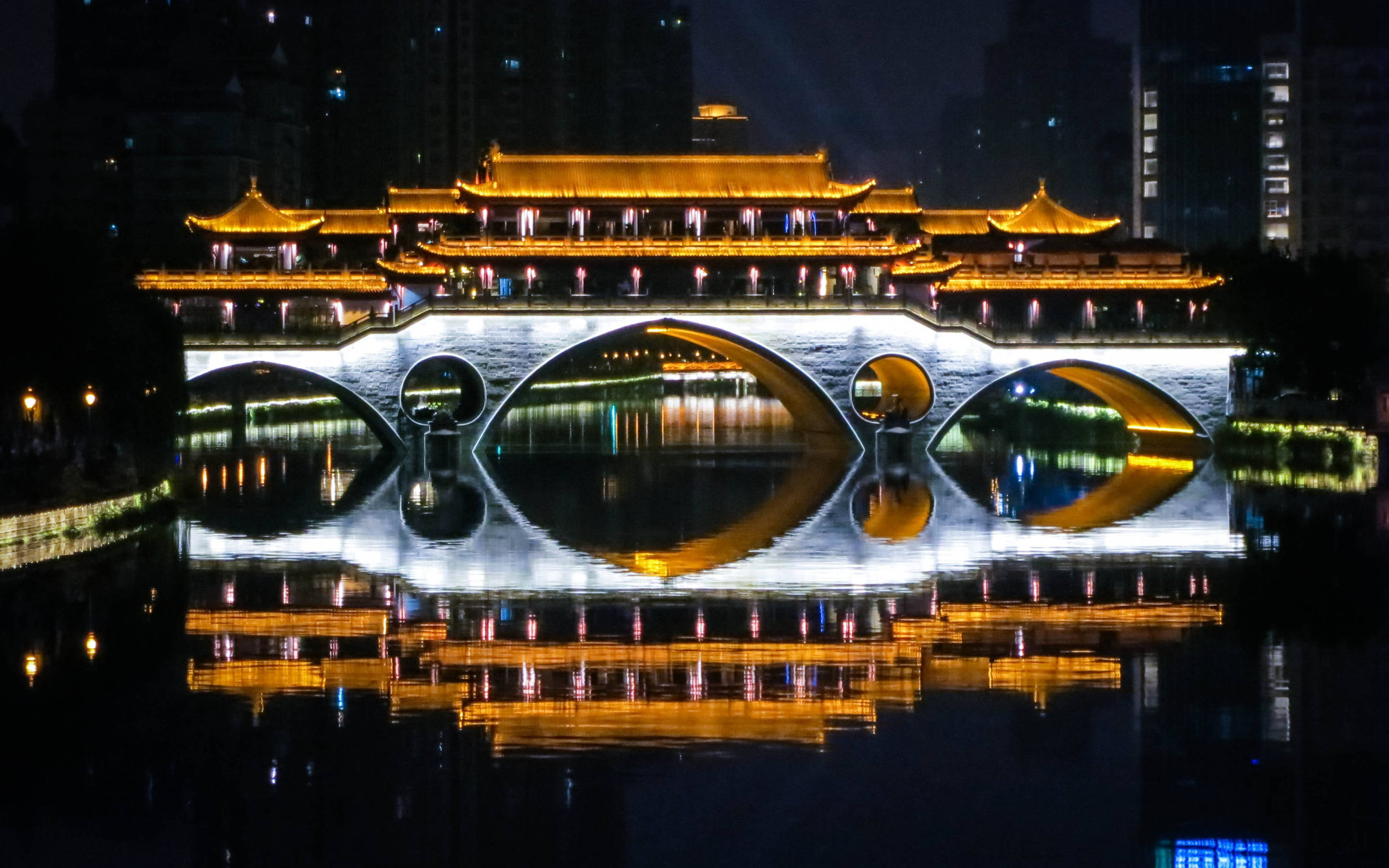 Chengdu 桌布 壁紙 — 一座宏偉的傳統中式橋樑，金色屋頂和白色拱門在夜晚平靜的水面上完美倒映，周圍環繞著城市燈光。