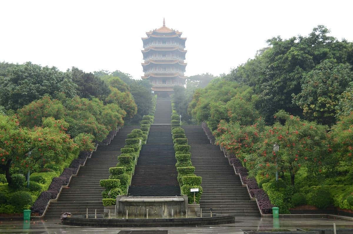 公園 桌布 壁紙 — 一座宏偉的傳統寶塔矗立在長長的石階頂端，兩旁是茂密的綠植和盛開的樹木，天空多雲。