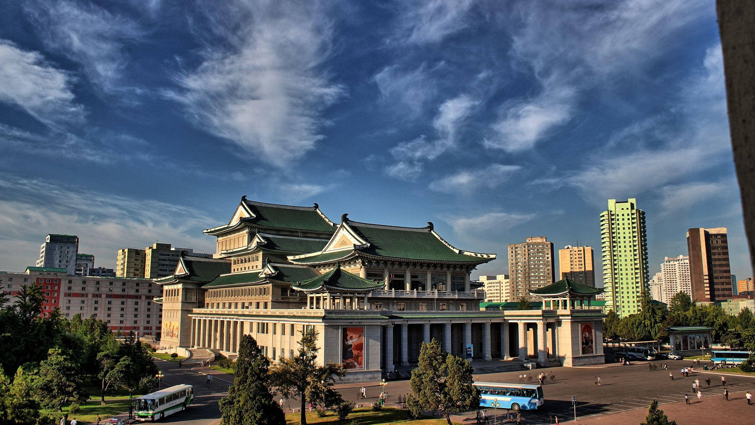 North Korea 桌布 壁紙 — 一座宏偉的傳統建築，擁有綠色屋頂，矗立在繁忙的城市廣場上，上方是帶有縷縷白雲的鮮豔藍天。