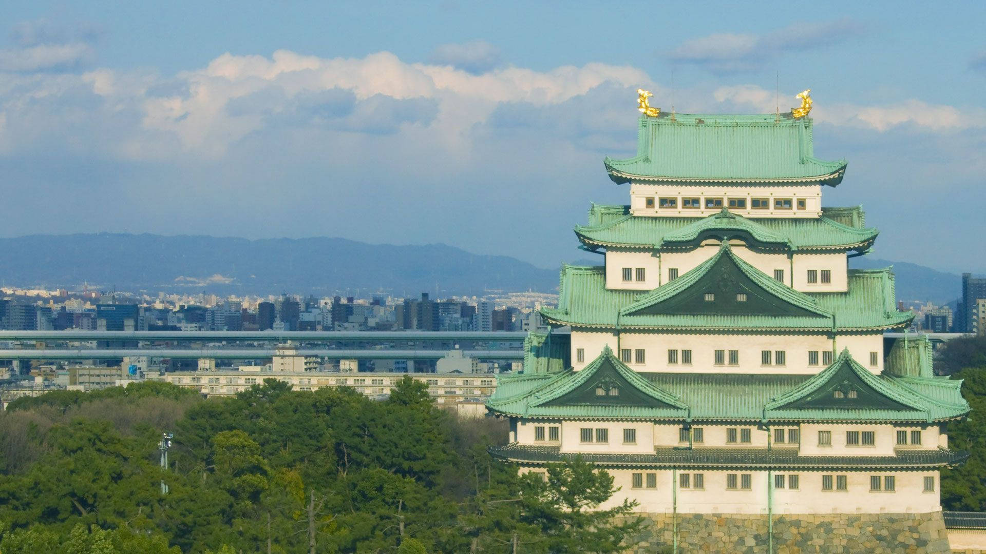 Nagoya 桌布 壁紙 — 一座宏偉的傳統日本城堡，擁有綠色瓦片屋頂，矗立在山丘上，俯瞰著一座龐大的現代城市和遠處的山脈，天空部分多雲。