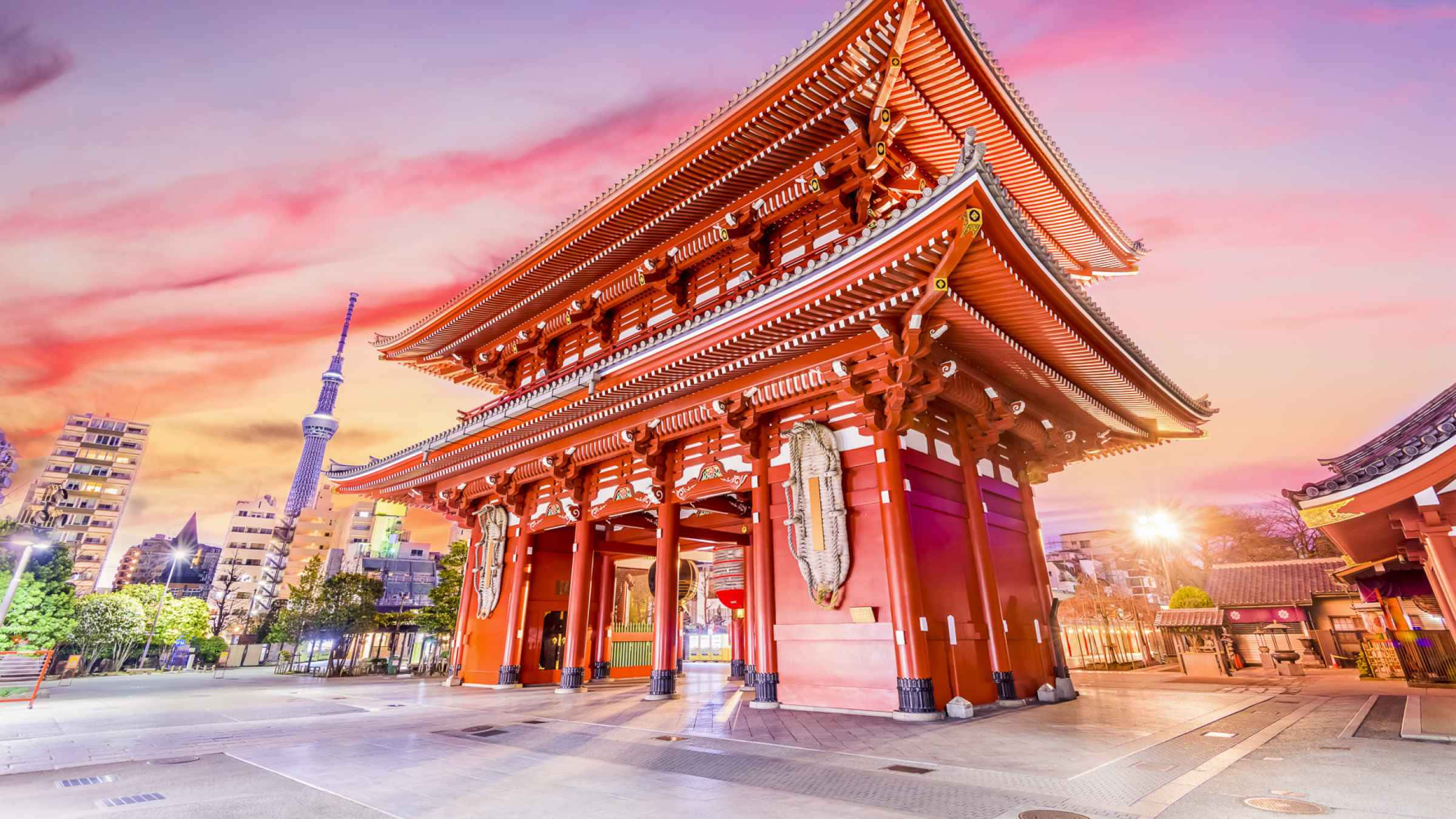 東京 桌布 壁紙 — 一座宏偉的傳統日本寺廟大門在溫暖的日落色調中閃耀，映襯著充滿活力的粉紅色和橙色天空，背景是現代城市天際線。