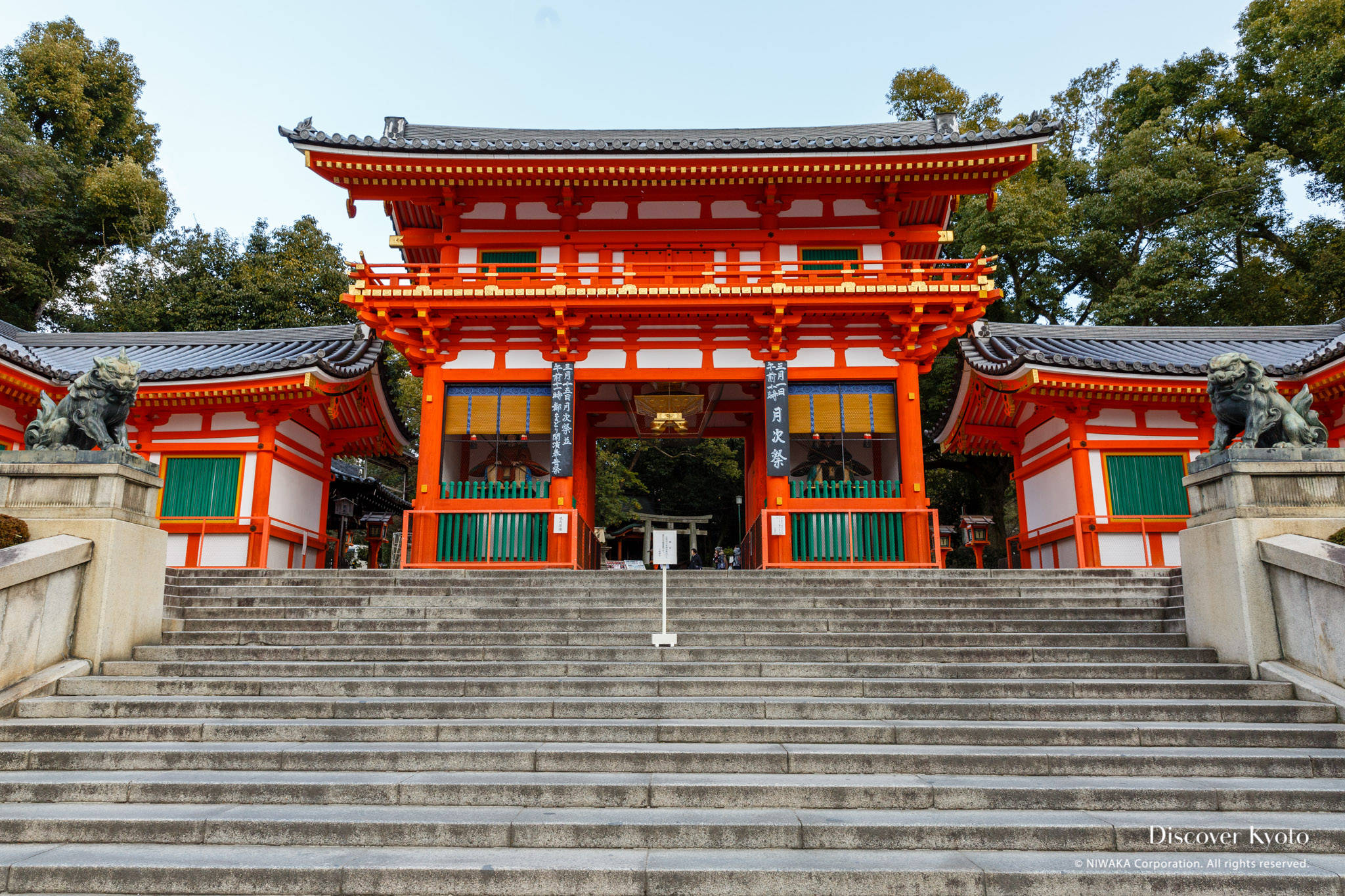 京都 桌布 壁紙 — 一座宏偉的傳統日本神社，擁有鮮豔的橙色鳥居和石階，兩側有守護獅子雕像，天空晴朗。