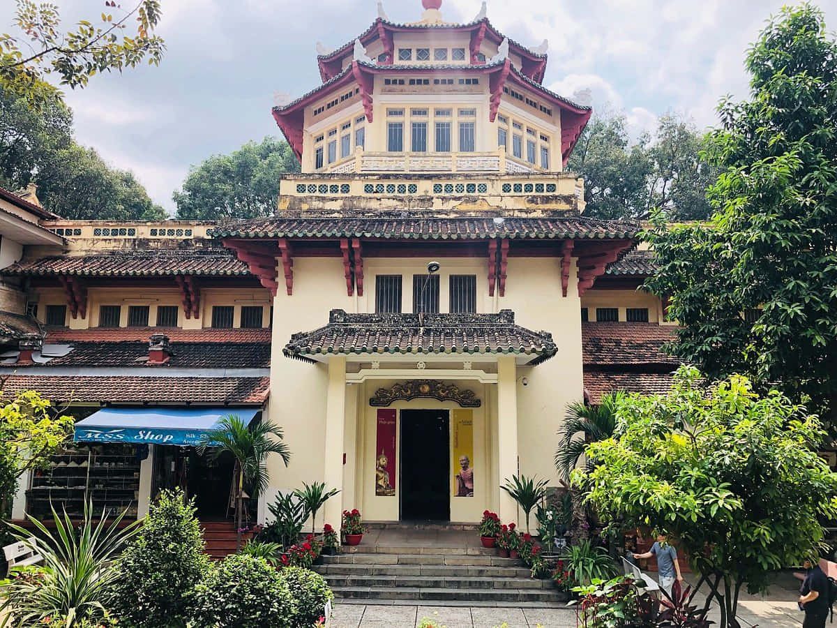 越南 桌布 壁紙 — 一座宏偉的傳統越南寺廟，擁有紅色分層屋頂和奶油色牆壁，矗立在晴朗天空下的茂密綠意之中。