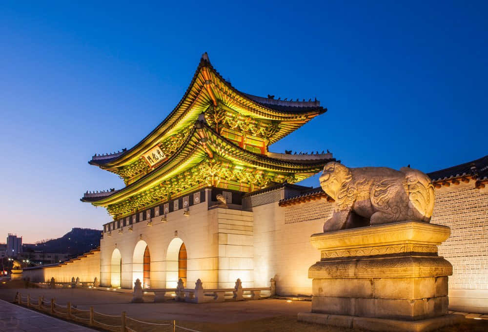 韓國 桌布 壁紙 — 一座宏偉的傳統韓國大門在黃昏時分被溫暖的燈光照亮，襯托著深藍色的天空，附近還有一座石獅守護雕像。