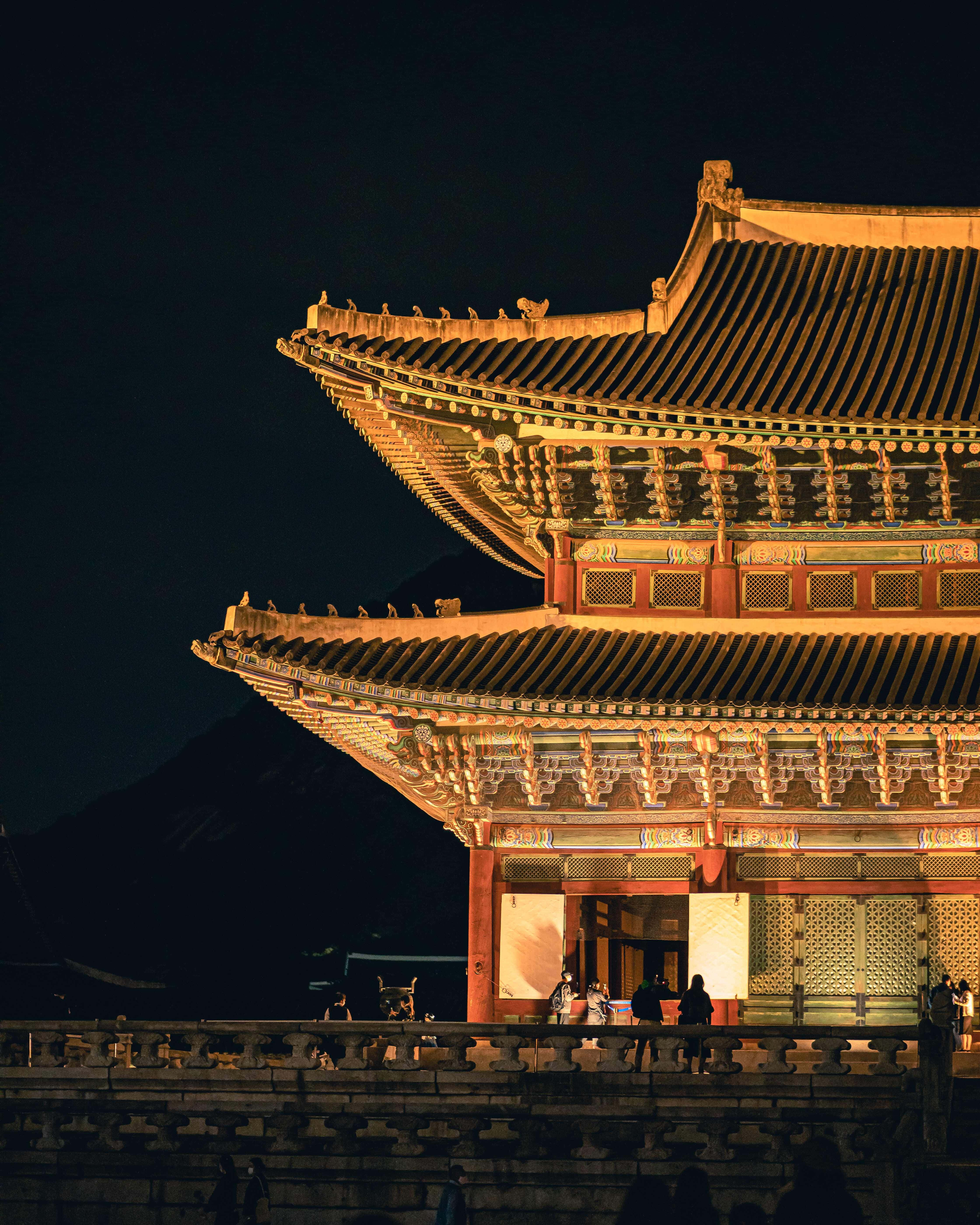 韓國 桌布 壁紙 — 一座宏偉的傳統韓國宮殿在夜晚以溫暖的金色燈光點亮，映襯著黑暗的天空。