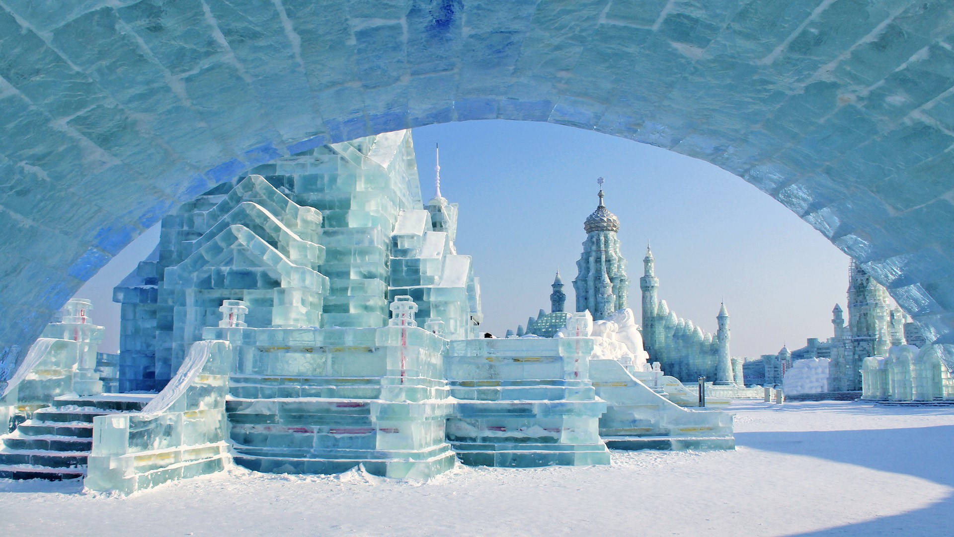 Harbin 桌布 壁紙 — 一座宏偉的冰城堡，擁有複雜的藍色冰拱門和塔樓，矗立在晴朗的天空下，置身於白雪皚皚的冬季仙境中。