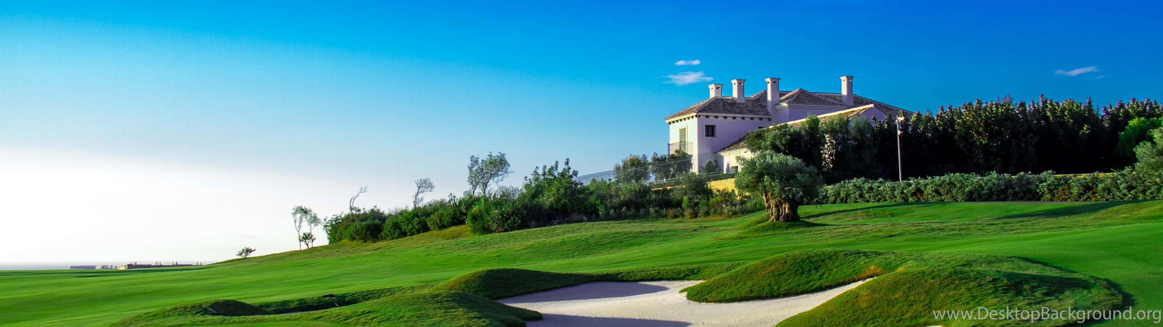 Golf 3840x1080 桌布 壁紙 — 一座宏偉的別墅坐落在鬱鬱蔥蔥的綠色山頂上，俯瞰著晴朗藍天下 pristine 的高爾夫球場。