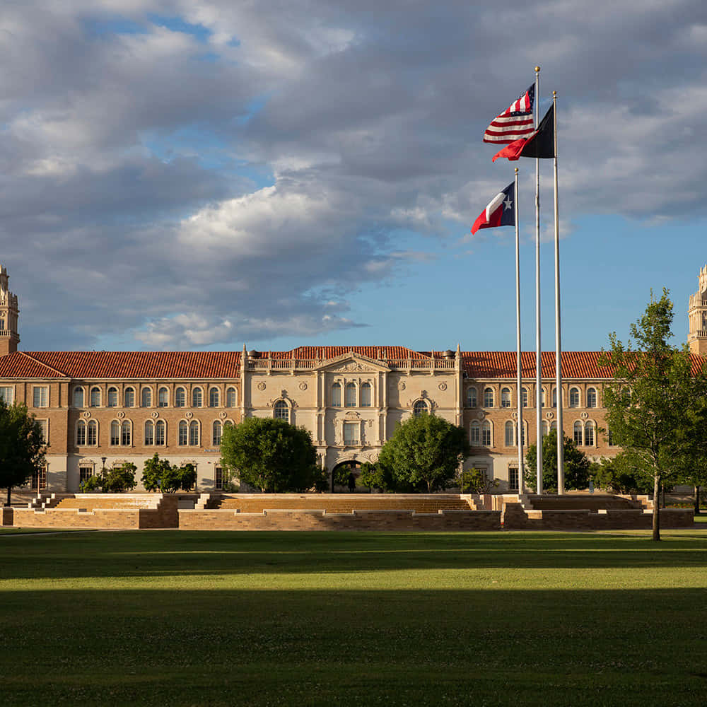 Texas Tech 桌布 壁紙 — 一座宏偉的古典大學建築，擁有紅色瓦片屋頂和三面飄揚的旗幟，矗立在部分多雲的天空下，前景是綠色草坪。