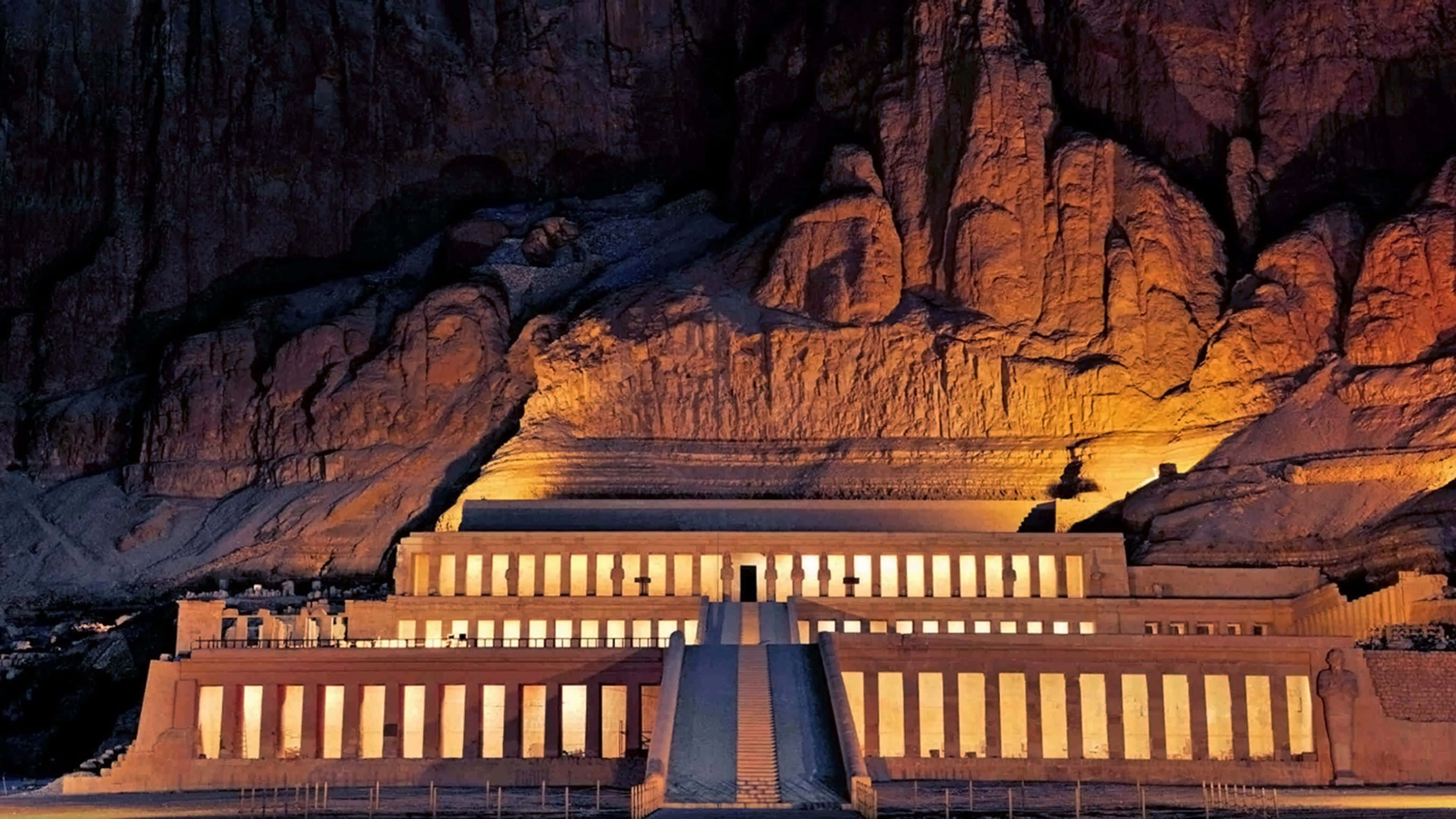 Ancient Egypt 桌布 壁紙 — 一座宏偉的古老寺廟雕刻在懸崖上，在夜間的人造燈光下溫暖地發光。