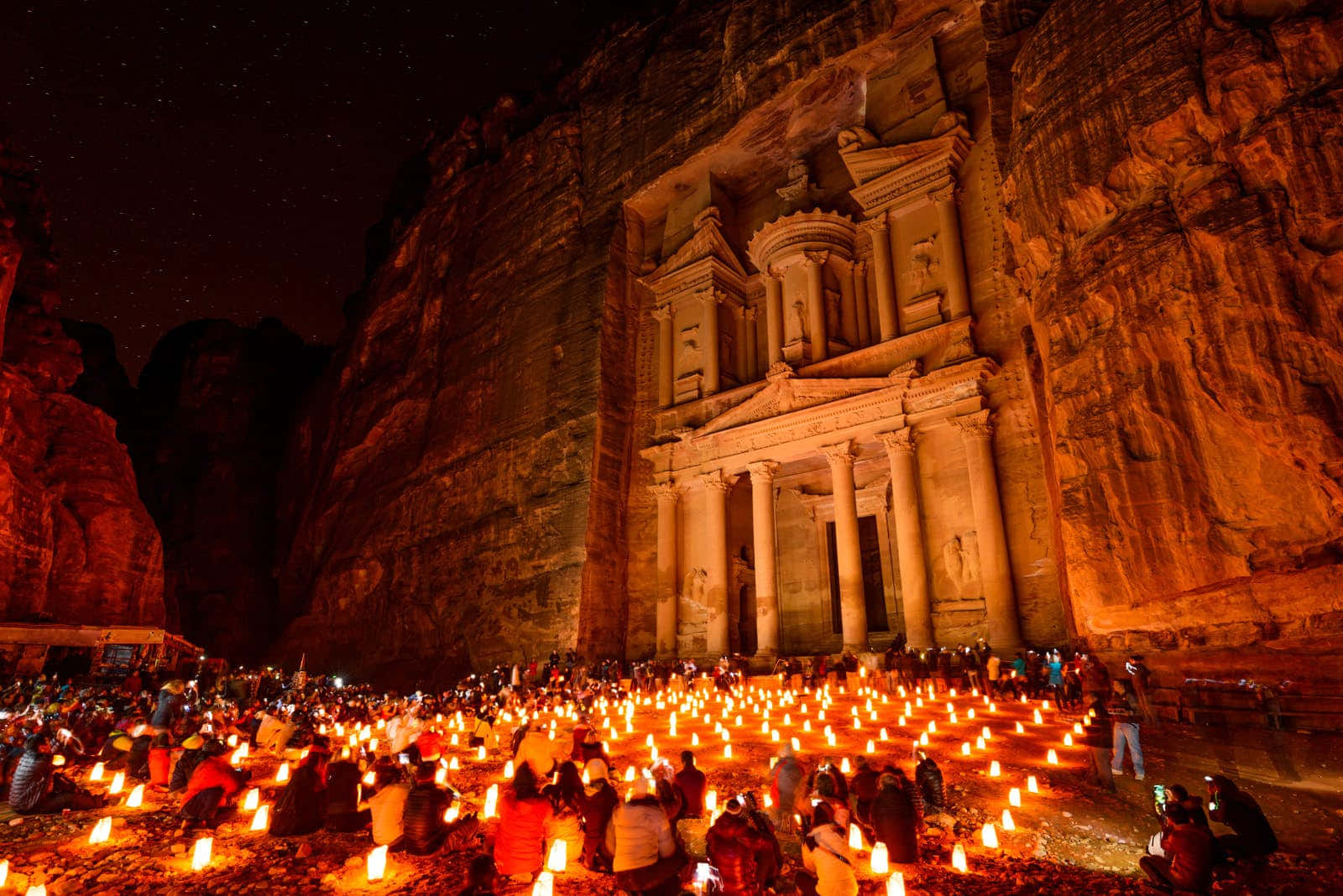 Jordan 桌布 壁紙 — 一座宏偉的古老寺廟雕刻在懸崖邊，在星空下溫暖地發光，數百人坐在發光的蠟燭中。