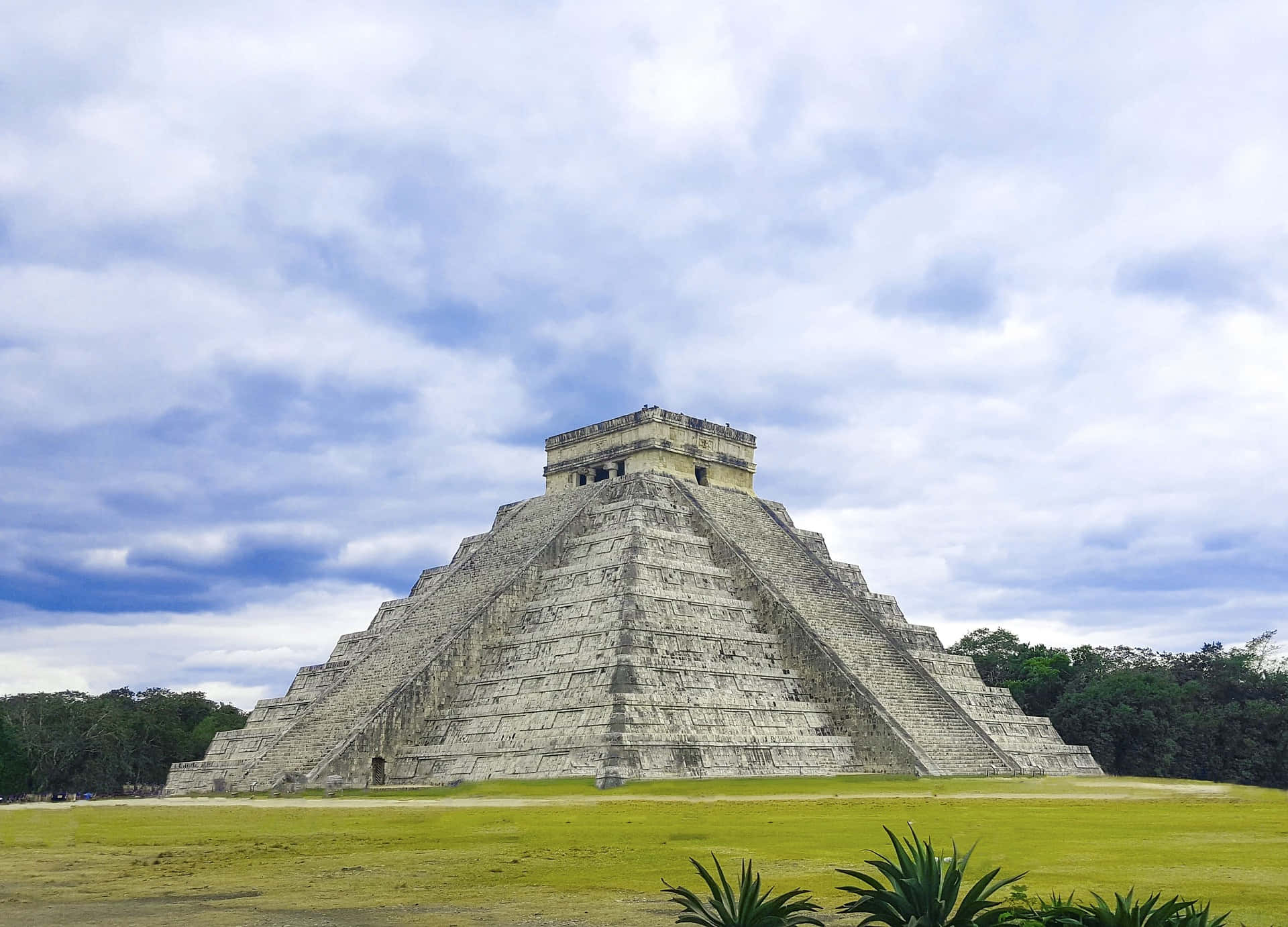 Cancun 桌布 壁紙 — 一座宏偉的古老瑪雅金字塔在陰雲密布的天空下巍然聳立，周圍環繞著茂密的綠草和熱帶植物。