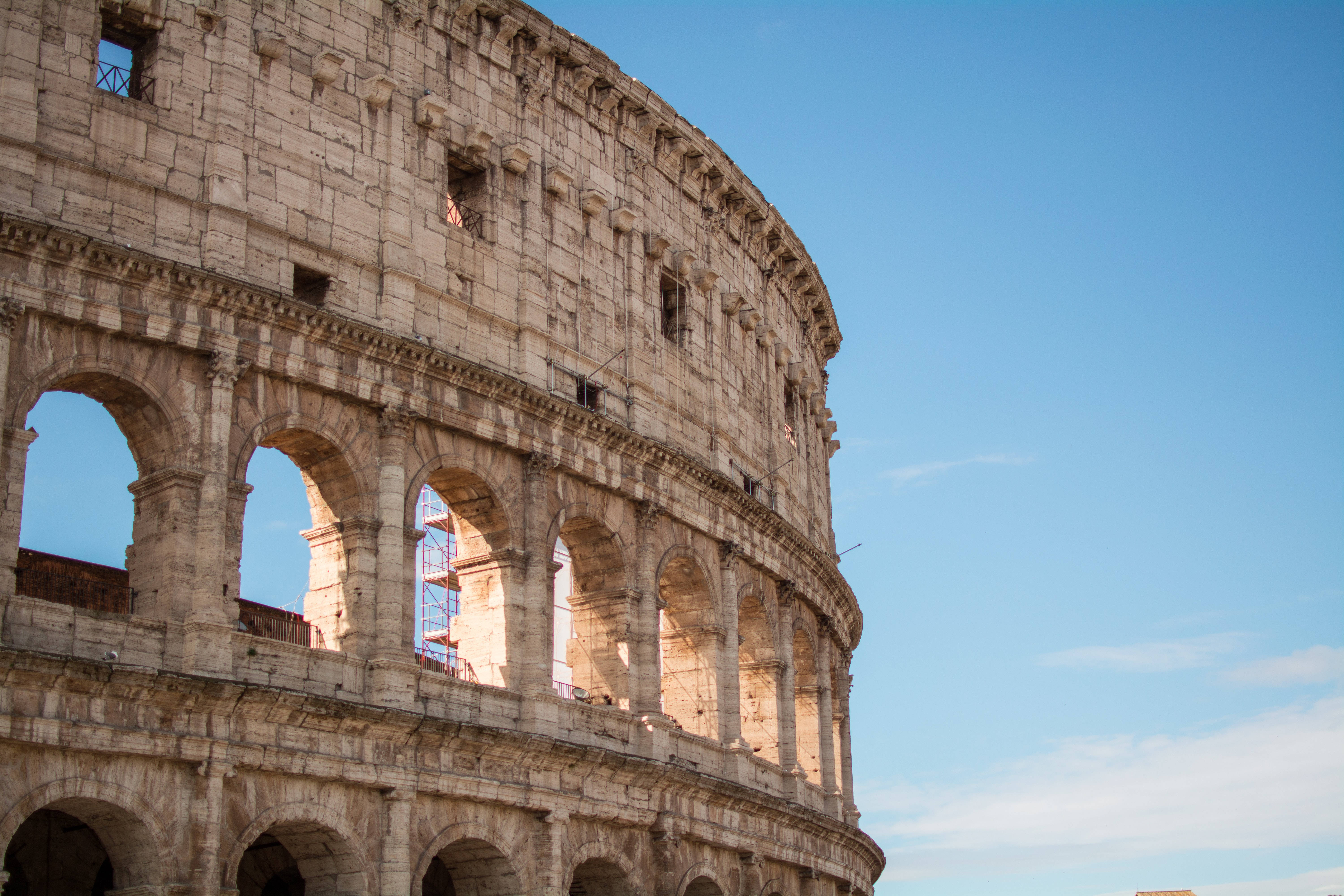 Rome 桌布 壁紙 — 一座宏偉的古老石造圓形競技場，帶有拱形開口，高聳於清澈的藍天下。