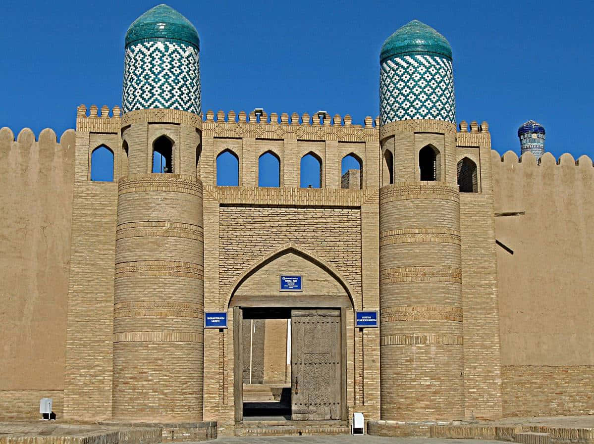 Uzbekistan 桌布 壁紙 — 一座宏偉的古老磚砌堡壘大門，在清澈的藍天下有兩個藍綠色瓷磚圓頂。