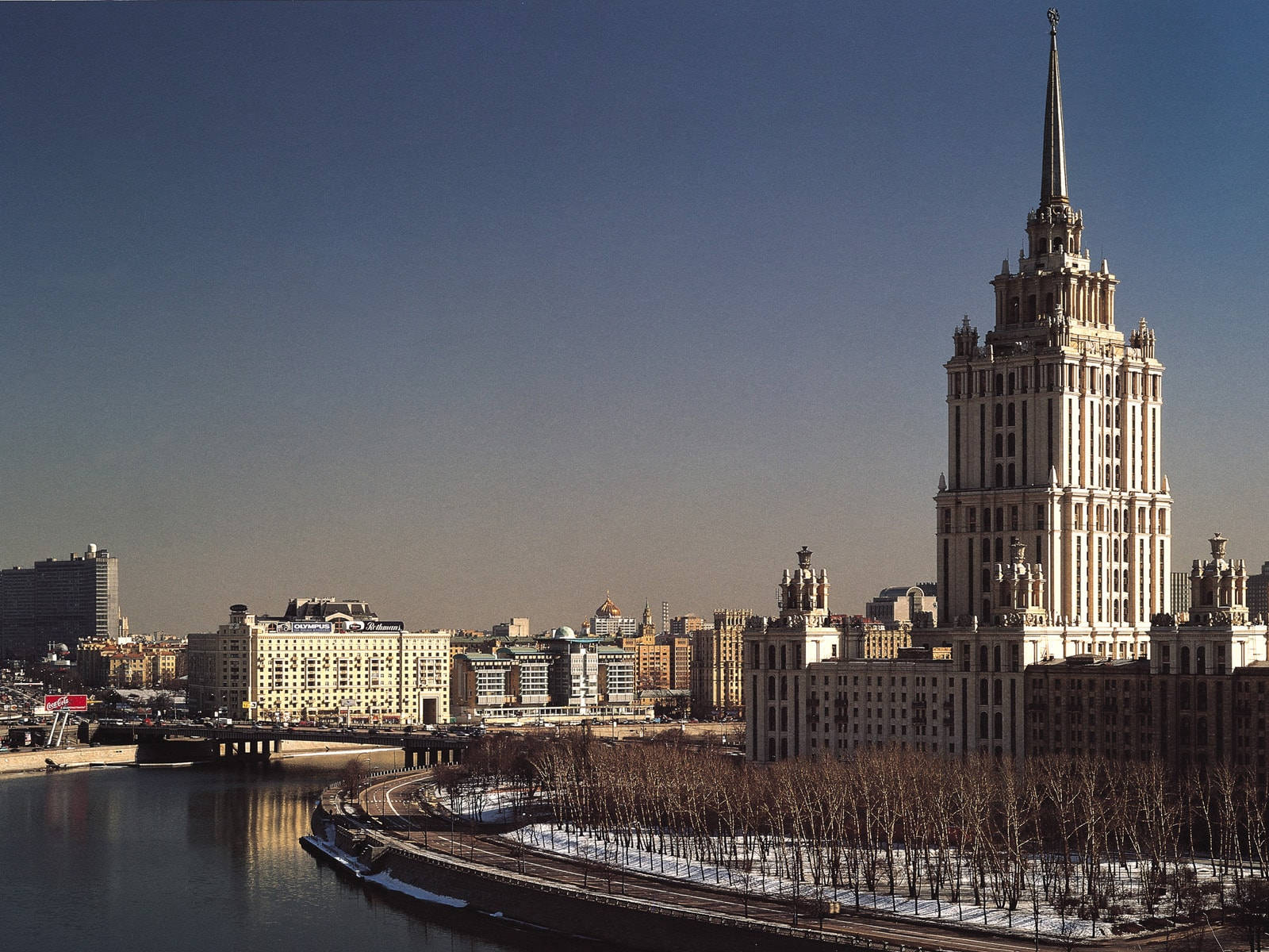 Moscow 桌布 壁紙 — 一座宏偉的史達林式摩天大樓主宰著冬日城市景觀，旁邊是一條覆蓋著薄雪的河流，在清澈的藍天下。