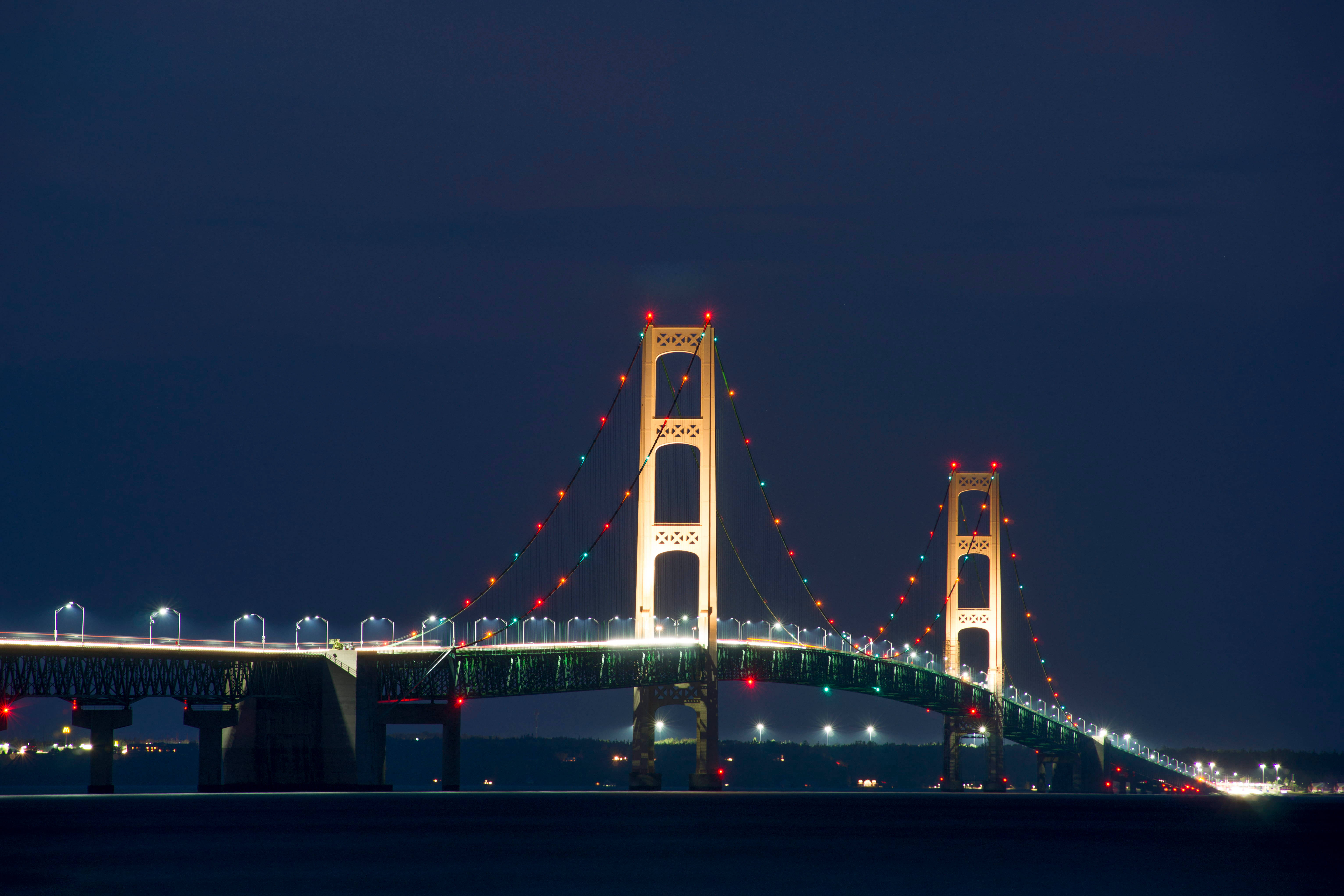 夜空 桌布 壁紙 — 一座宏偉的吊橋在平靜的水面上，以深沉的暮色天空為背景，閃爍著節日燈光。