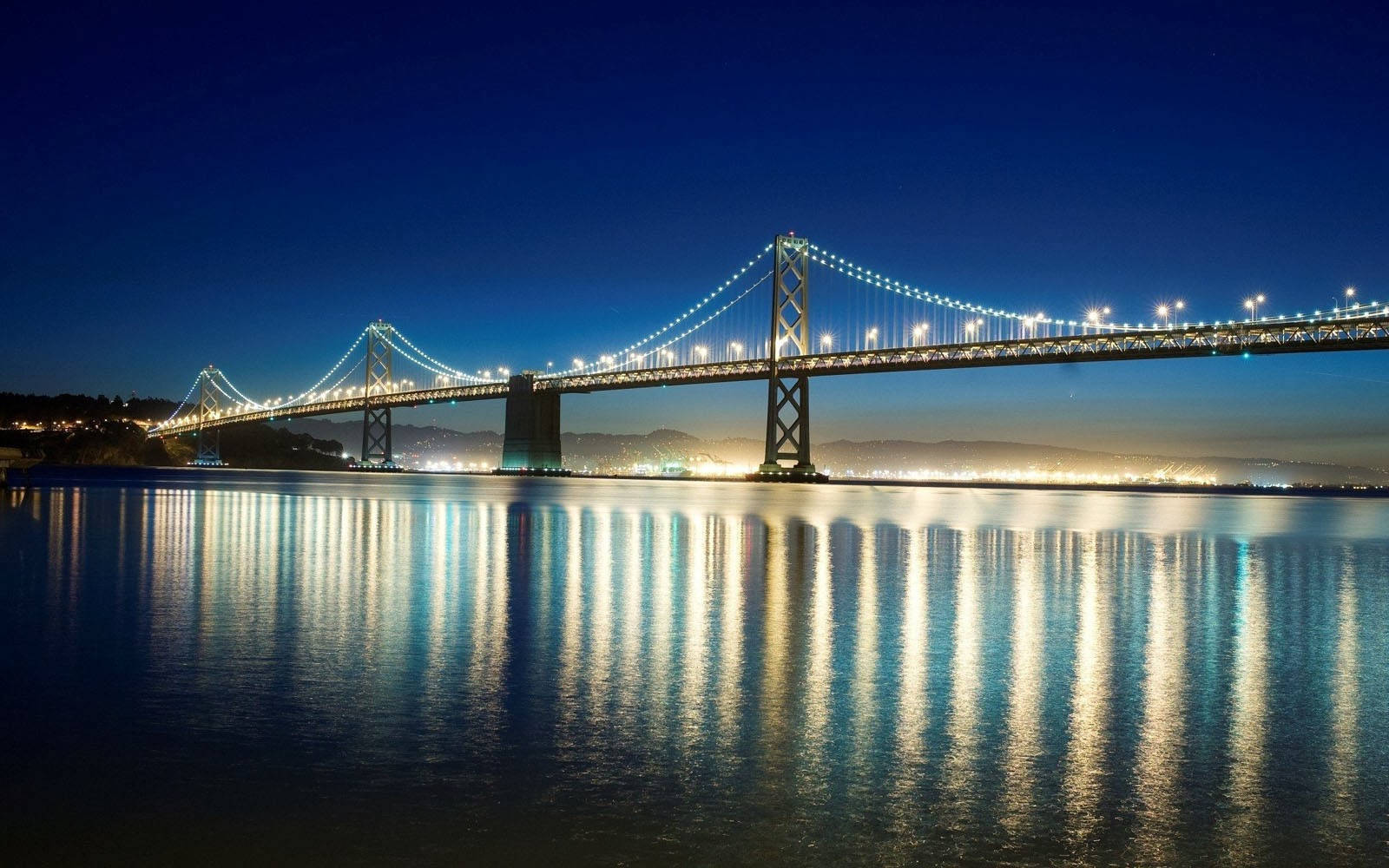 San Francisco HD 桌布 壁紙 — 一座宏偉的吊橋在深藍色的夜空下散發著溫暖的光芒，其倒影在下方平靜的水面上閃爍。