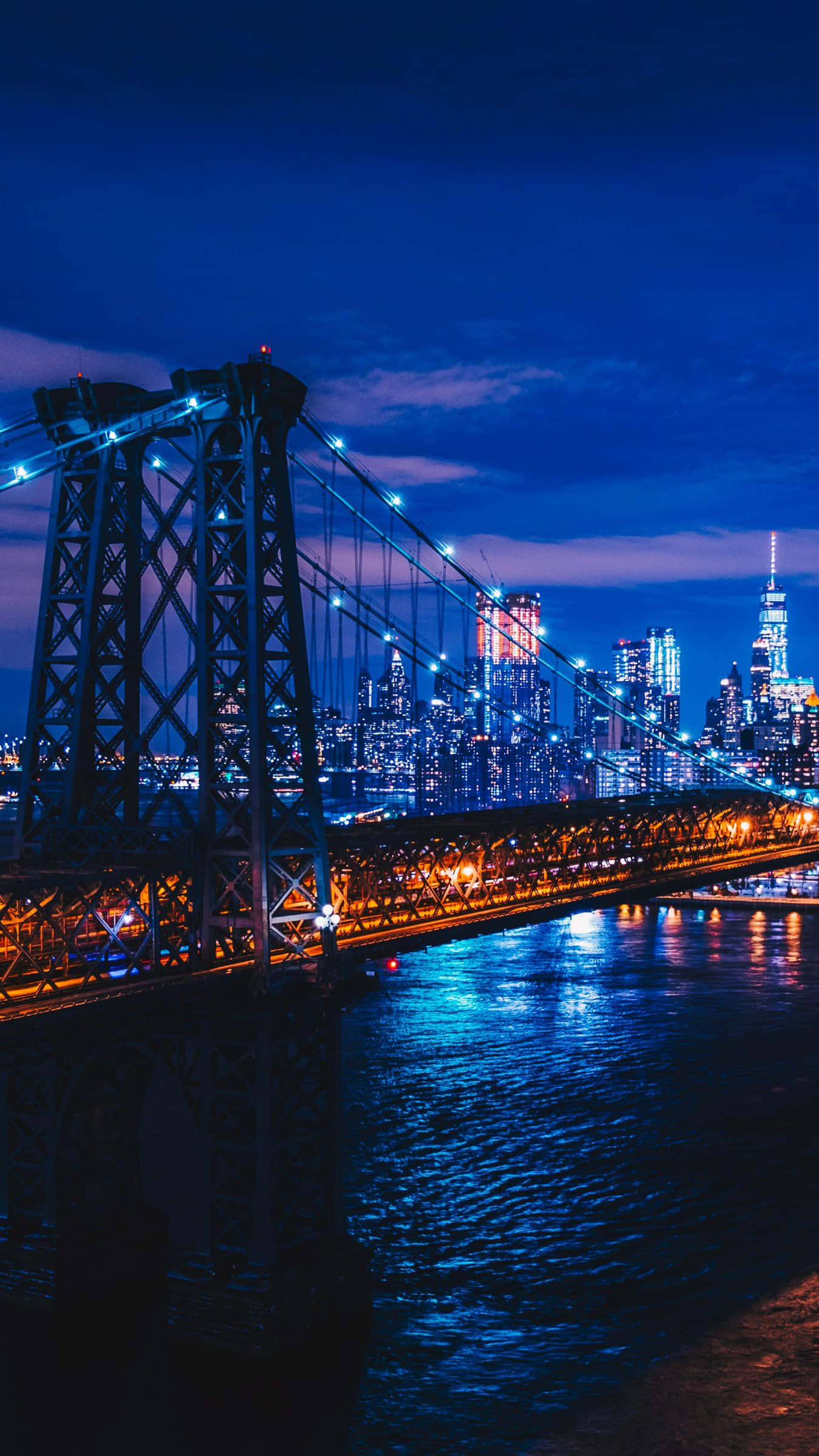 New York City Night iPhone 桌布 壁紙 — 一座宏偉的吊橋在深藍色的暮色天空下散發著溫暖的光芒，倒映在下面的水面上，遠處的城市天際線閃爍著。