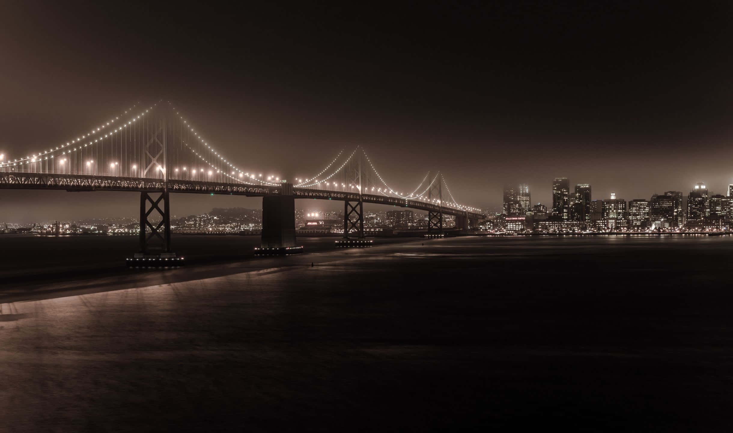 2440x1440 San Francisco 桌布 壁紙 — 一座宏偉的吊橋在漆黑的夜空下閃爍著溫暖的燈光，橫跨平靜的水道，通往遠處的城市天際線。