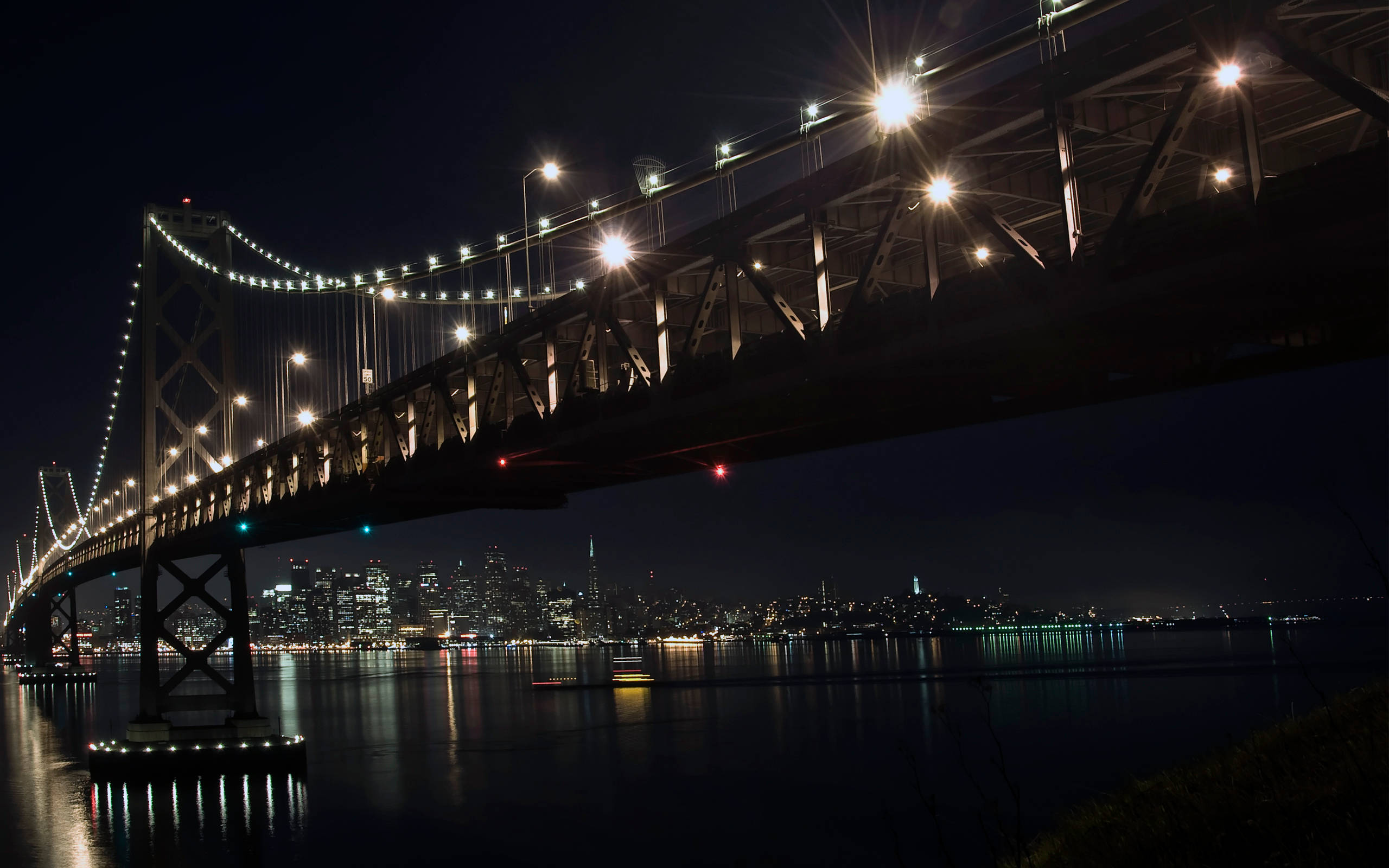 lights 桌布 壁紙 — 一座宏偉的吊橋在漆黑的夜空中散發著溫暖的光芒，其倒影在下方平靜的水面上閃爍。