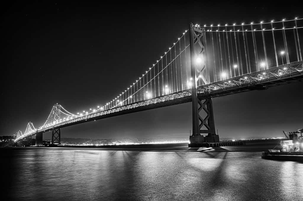 San Francisco Black And White 桌布 壁紙 — 一座宏偉的吊橋在繁星點點的夜空下閃耀著明亮的燈光，其倒影在下方平靜的水面上閃爍。