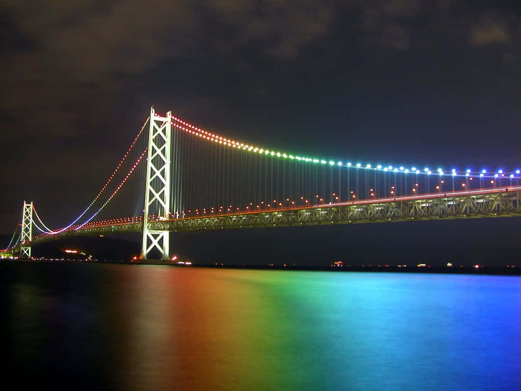 Rainbow Bridge 桌布 壁紙 — 一座宏偉的吊橋在陰暗的夜空下，以彩虹燈光橫跨黑暗的水道。