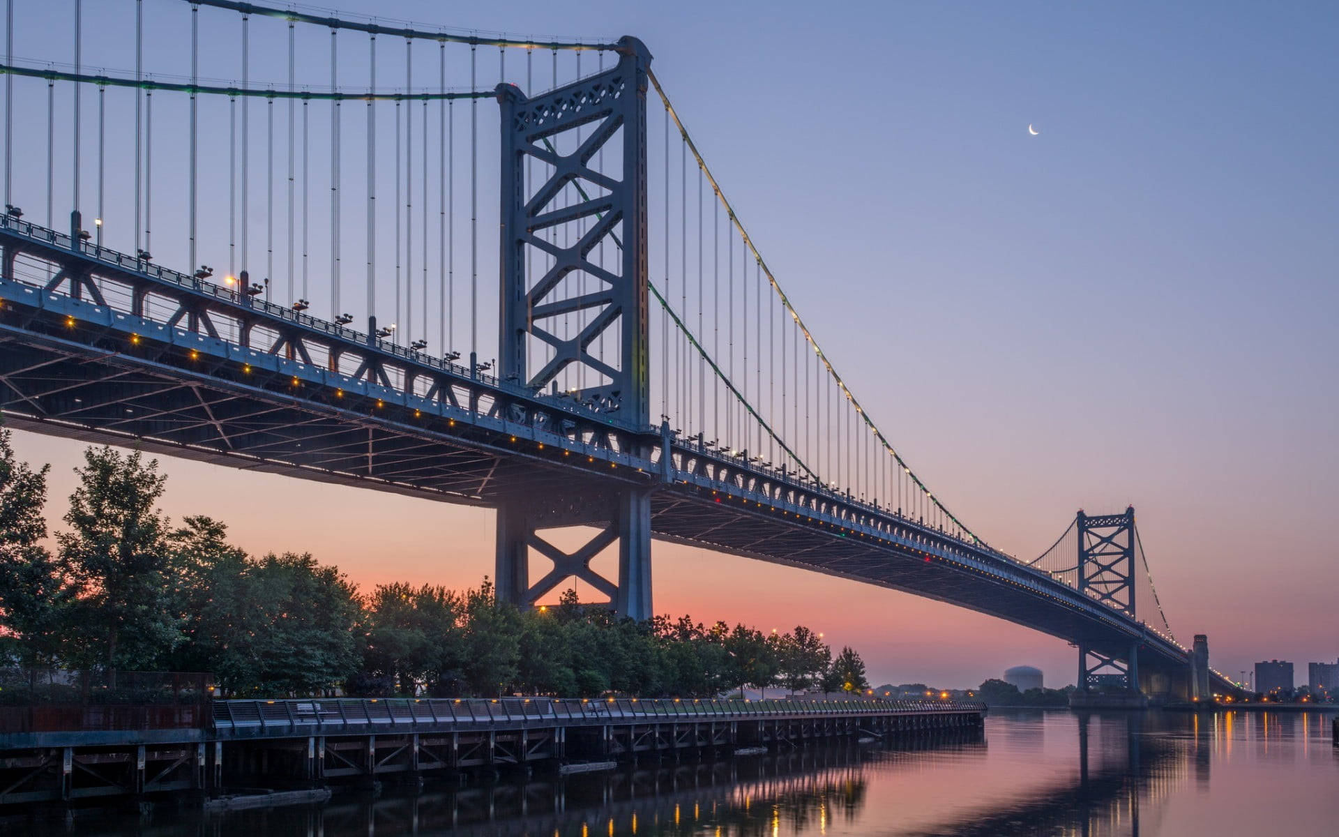 Philadelphia 桌布 壁紙 — 一座宏偉的吊橋橫跨在黃昏時平靜的河面上，上方有一彎新月，溫暖的燈光倒映在水中。