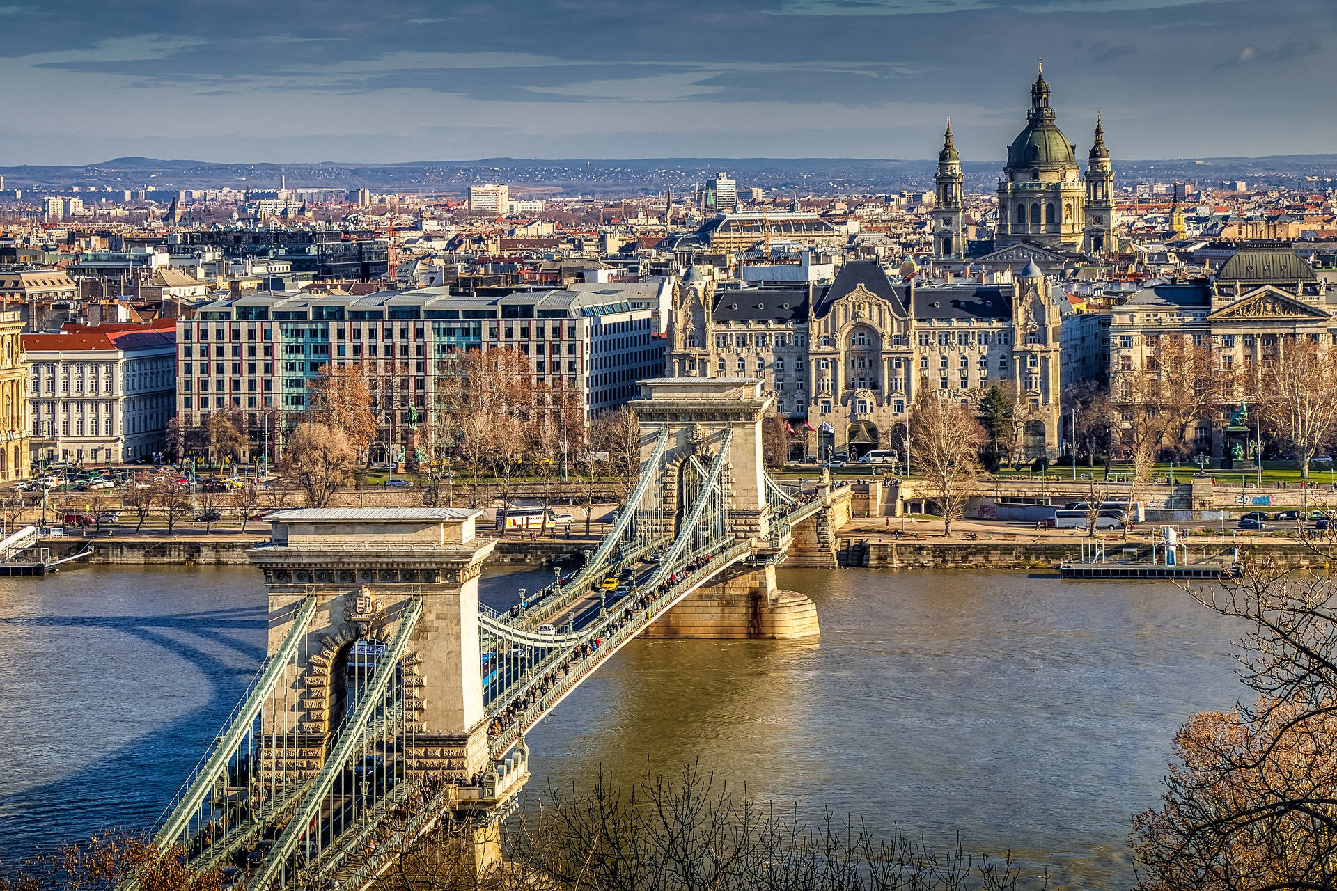 budapest 桌布 壁紙 — 一座宏偉的吊橋橫跨寬闊的河流，天空多雲，背景是歷史建築和一座圓頂大教堂。