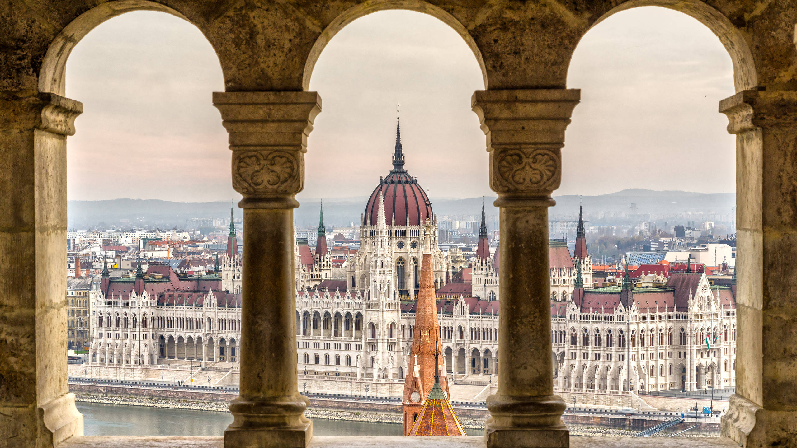 budapest 桌布 壁紙 — 一座宏偉的哥德式國會大廈，擁有紅色屋頂和尖塔，透過風化的石拱門俯瞰河流。