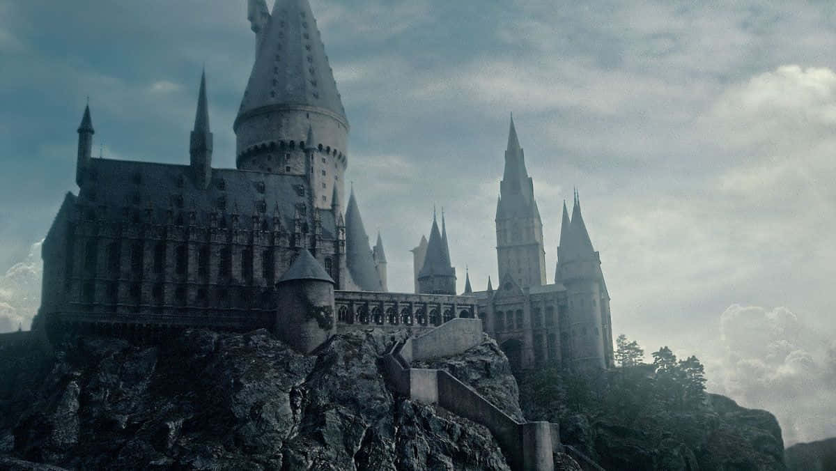 Hogwarts Castle 桌布 壁紙 — 一座宏偉的哥德式城堡，擁有尖塔和角樓，坐落在多雲天空下的岩石懸崖頂上。