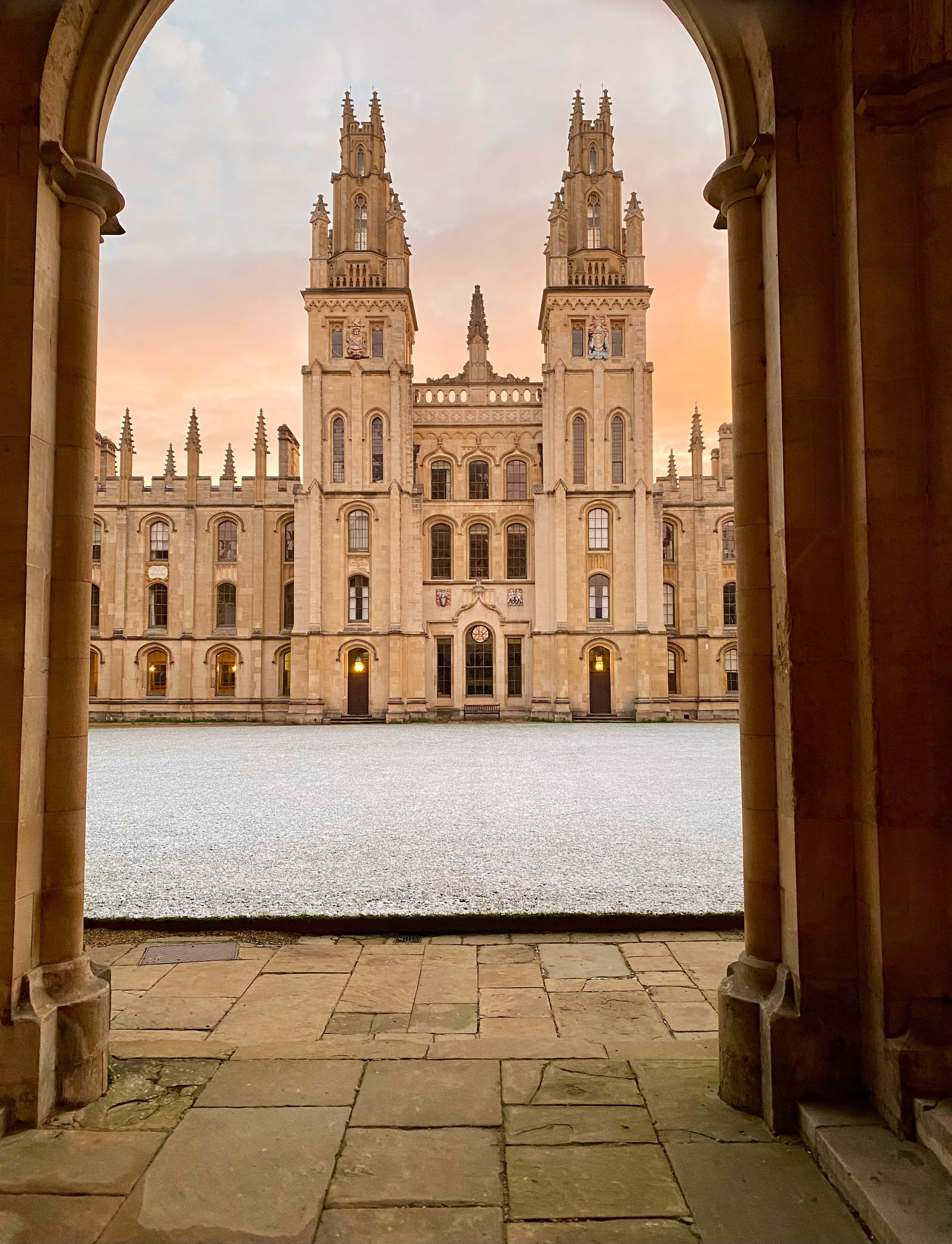 England 桌布 壁紙 — 一座宏偉的哥德式大學建築，擁有雙塔，倒映在平靜的水面上，在日落時分由拱形石門框住。