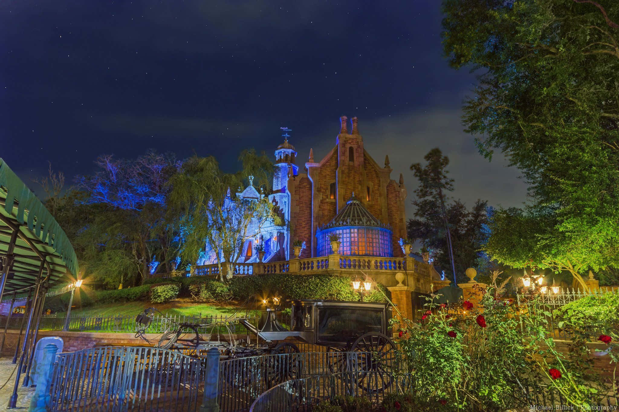 Disney World Desktop 桌布 壁紙 — 一座宏偉的哥德式大宅，在星空下以藍色燈光照明，周圍環繞著茂盛的花園和復古馬車。