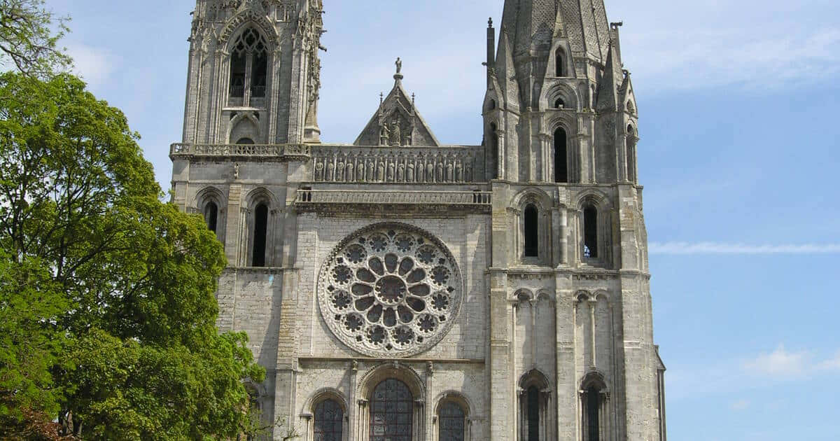 Gothic Architecture 桌布 壁紙 — 一座宏偉的哥德式大教堂，擁有雙石塔和一個大型玫瑰窗，在晴朗的藍天和附近茂密的綠樹襯托下巍然聳立。