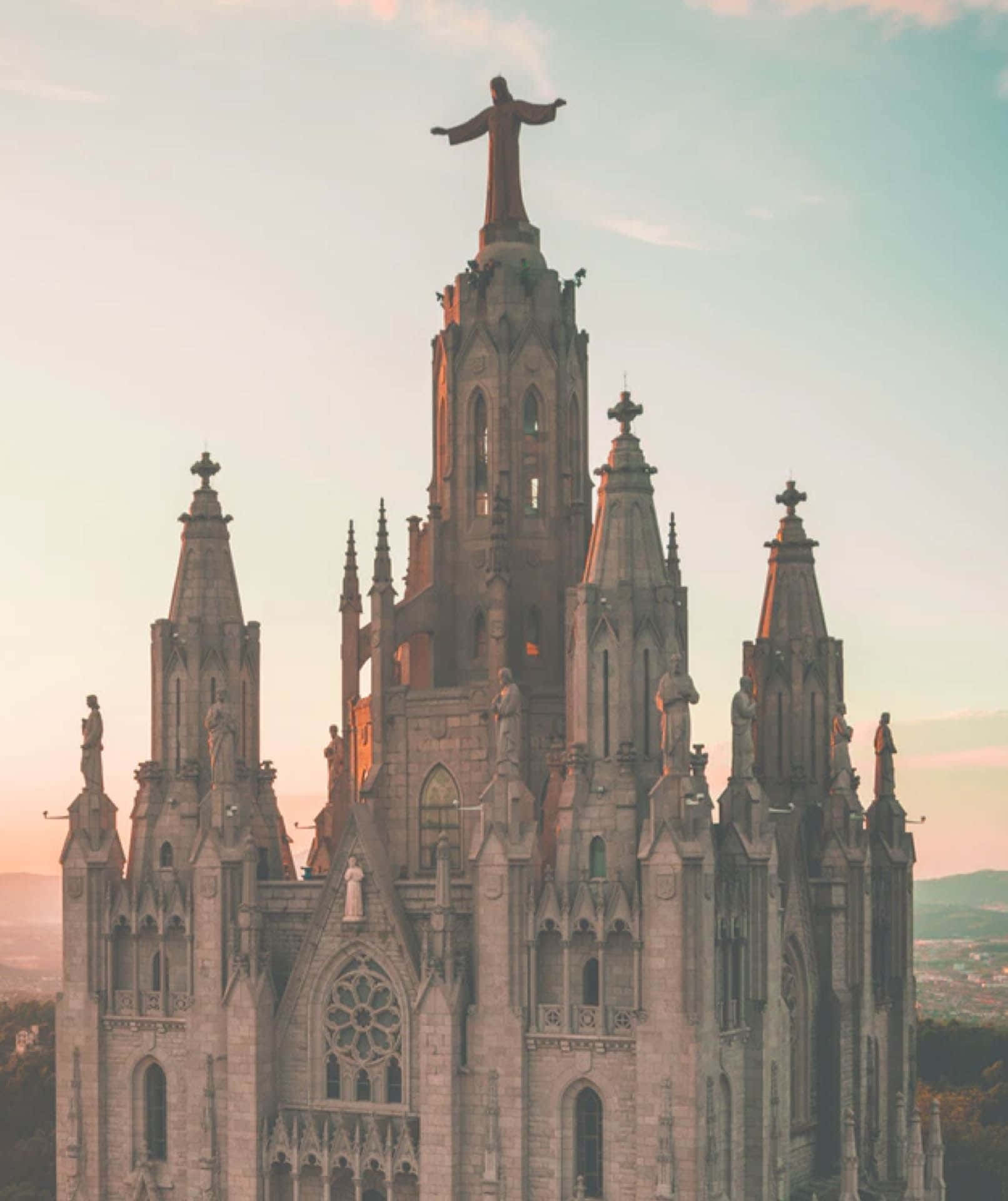 Barcelona 桌布 壁紙 — 一座宏偉的哥德式大教堂，矗立著高大的基督雕像，在日落時分將溫暖的金色光芒灑在其石質尖塔上。