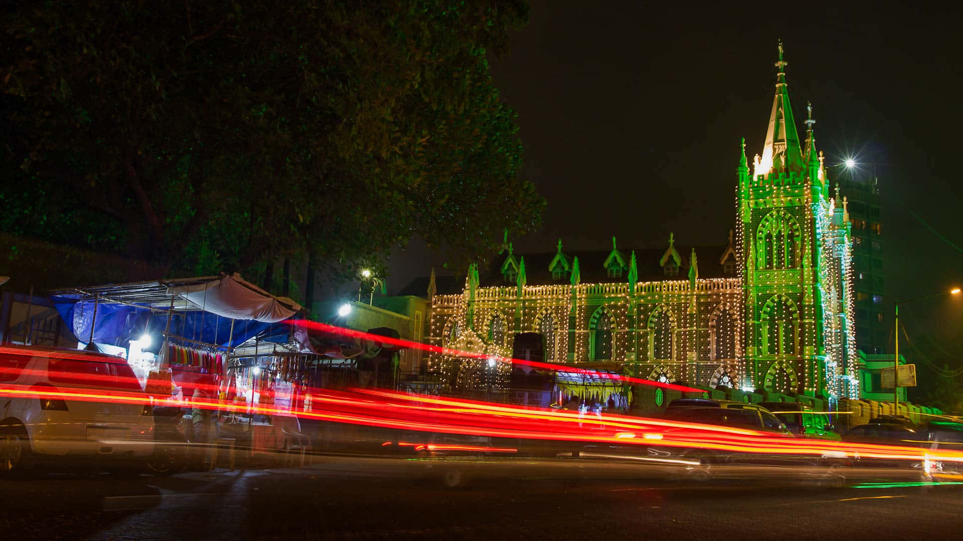 Mumbai 桌布 壁紙 — 一座宏偉的哥德式教堂在夜間被綠色燈光照亮，前景街道上紅色車燈的條紋模糊掠過。