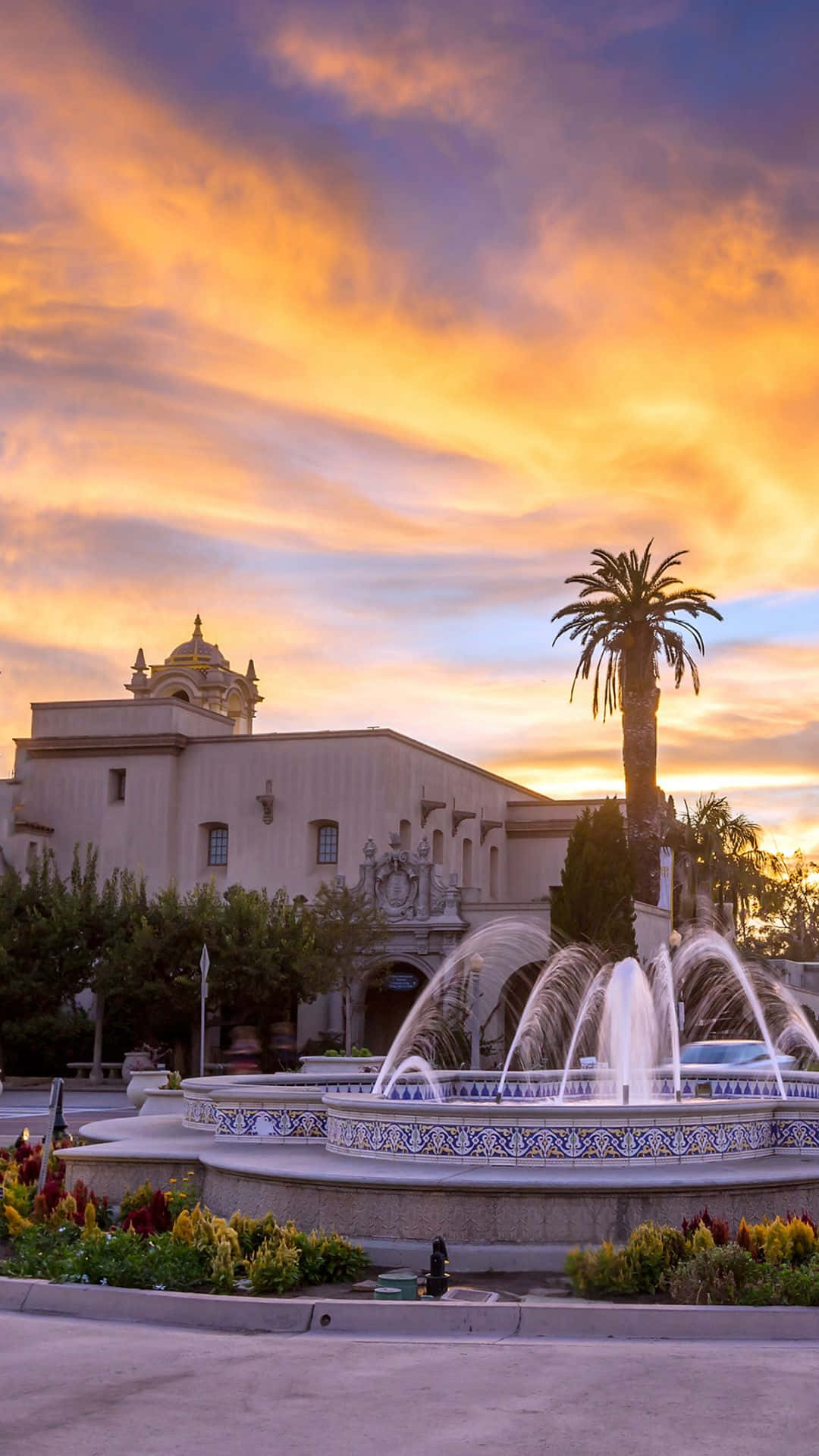 San Diego iPhone 桌布 壁紙 — 一座宏偉的噴泉在火紅的日落天空下，於歷史建築和棕櫚樹上方噴出水柱。
