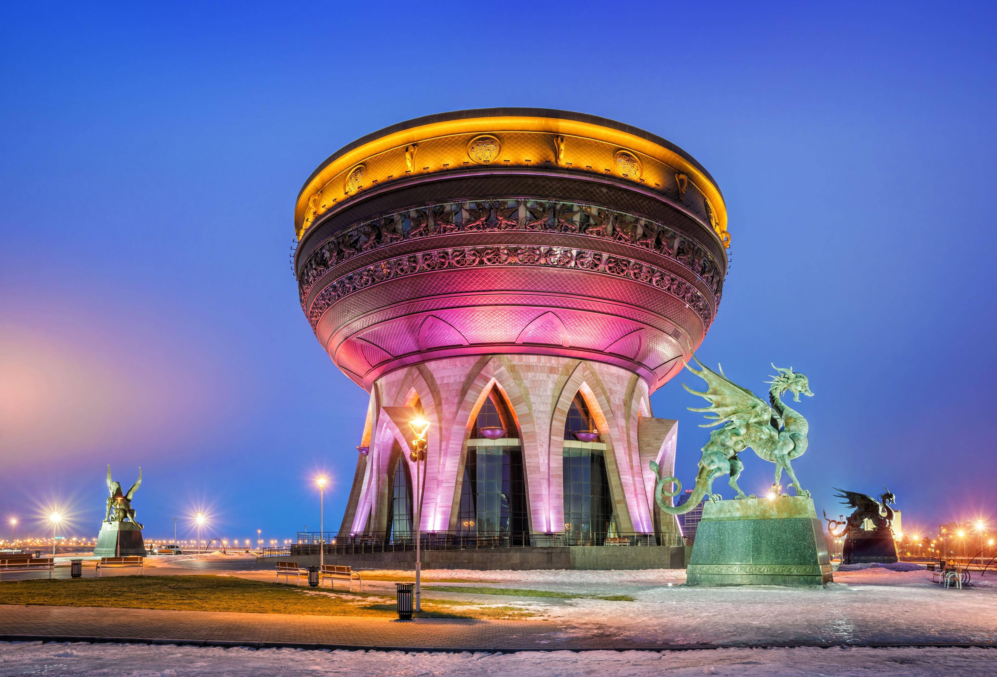 Kazan 桌布 壁紙 — 一座宏偉的圓形建築物亮起燈光，龍雕像矗立在雪中，背景是暮色天空。
