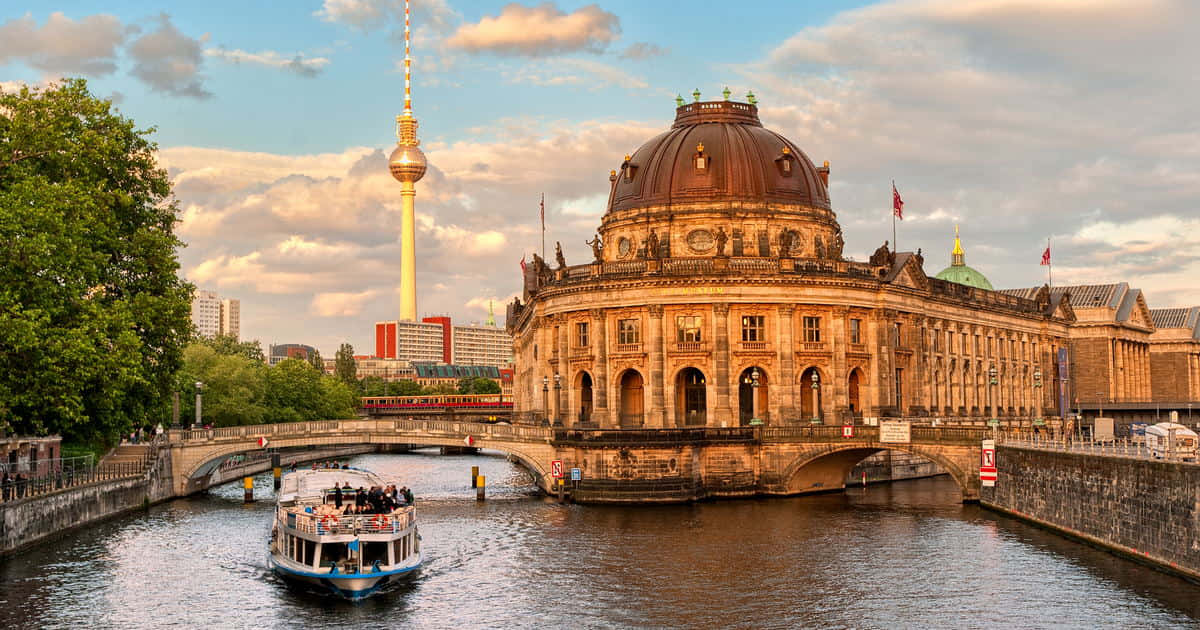 Berlin 桌布 壁紙 — 一座宏偉的圓頂建築和標誌性電視塔矗立在河邊，一艘觀光船在黃金時段從橋下穿過。