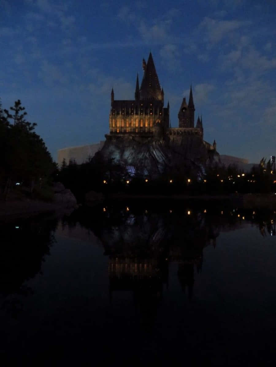 The Hogwarts Lake 桌布 壁紙 — 一座宏偉的城堡，窗戶閃爍著光芒，在暮色天空下倒映在平靜的水面上。