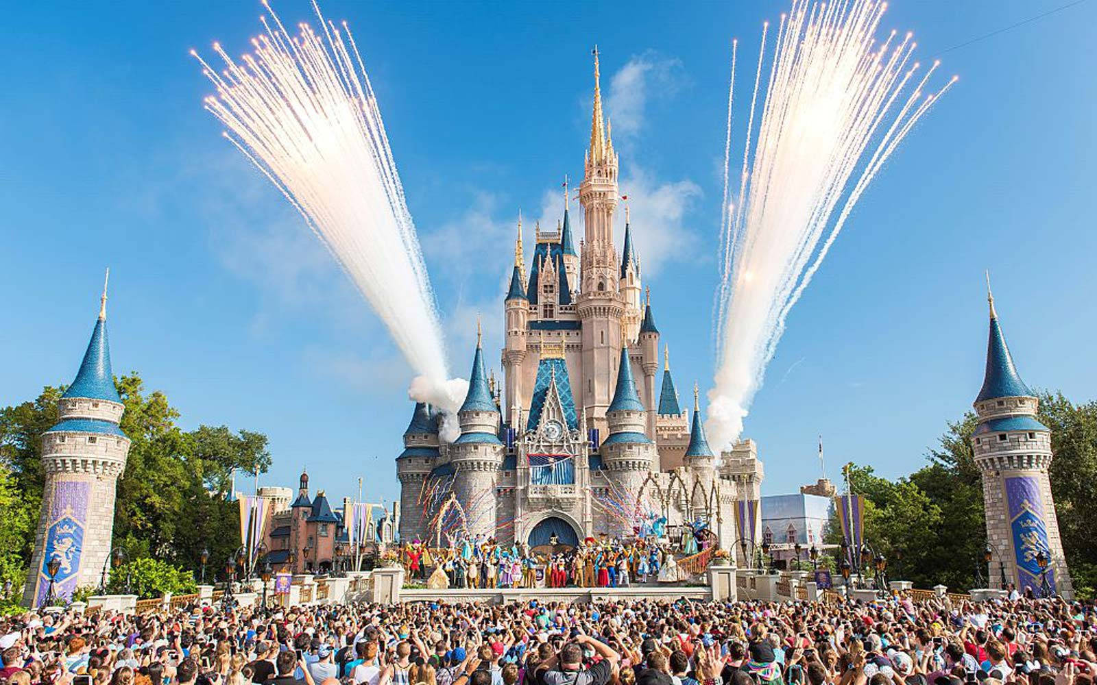 Disneyland Castle 桌布 壁紙 — 一座宏偉的城堡，藍色塔樓在絢麗的煙火中綻放，歡呼的人群在晴朗的藍天下觀看。