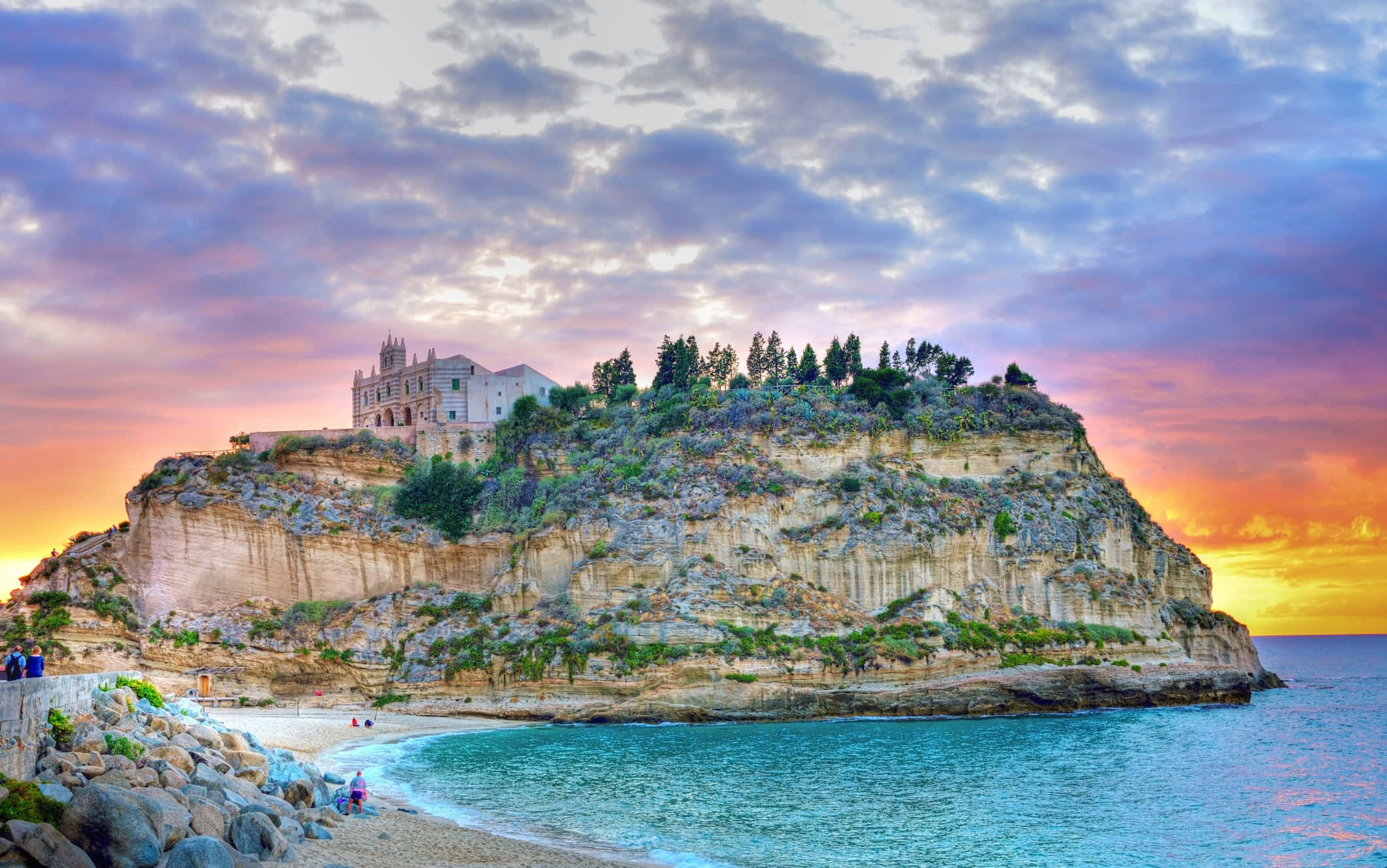Italian Beach 桌布 壁紙 — 一座宏偉的城堡坐落在崎嶇的懸崖頂上，俯瞰著日落時分碧綠色的海洋，天空呈現出鮮豔的紫色和橙色。