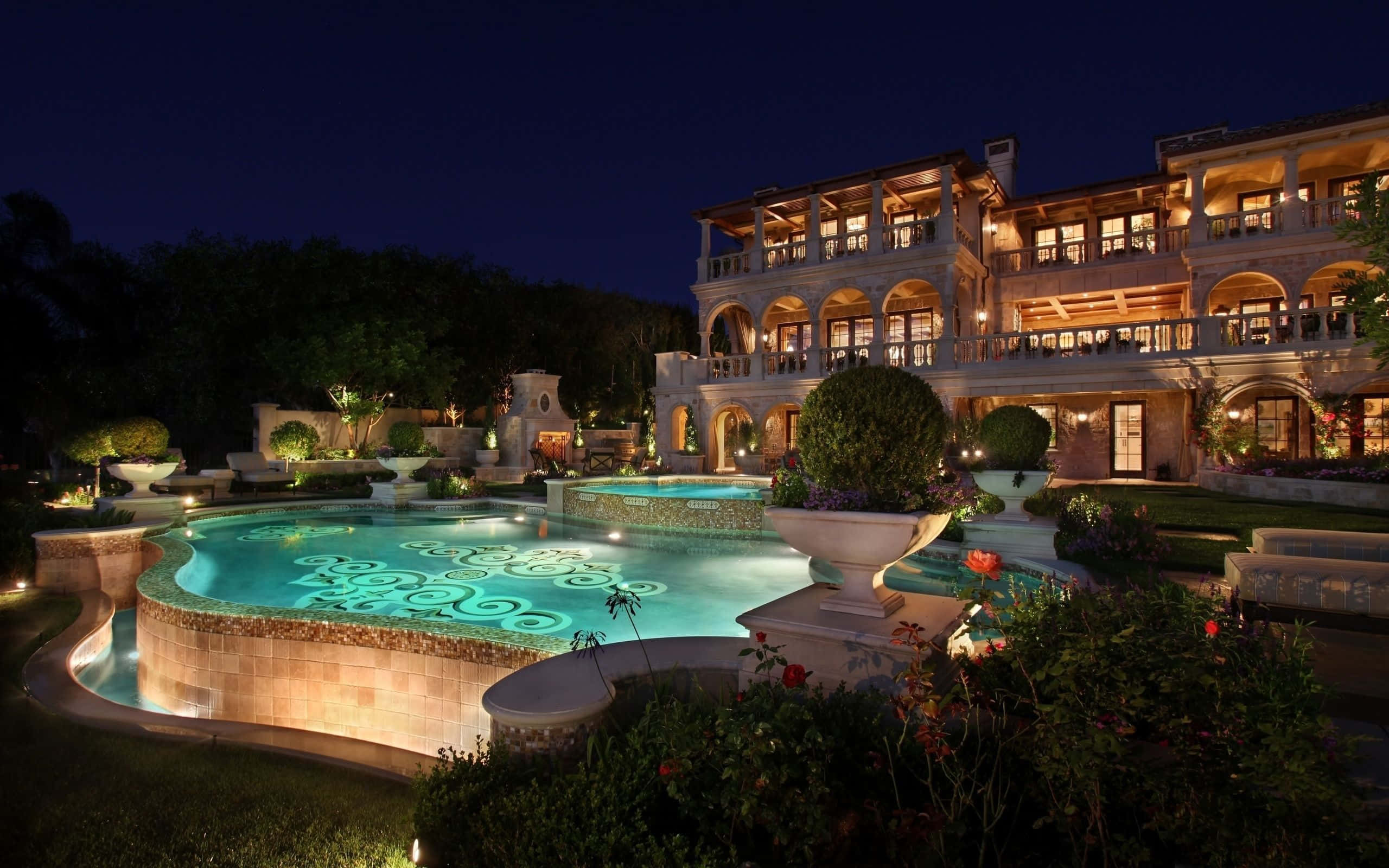 Mansion 桌布 壁紙 — 一座宏偉的宅邸在夜間亮起燈光，設有發光的游泳池，呈現旋轉的光影圖案，周圍環繞著修剪整齊的花園。