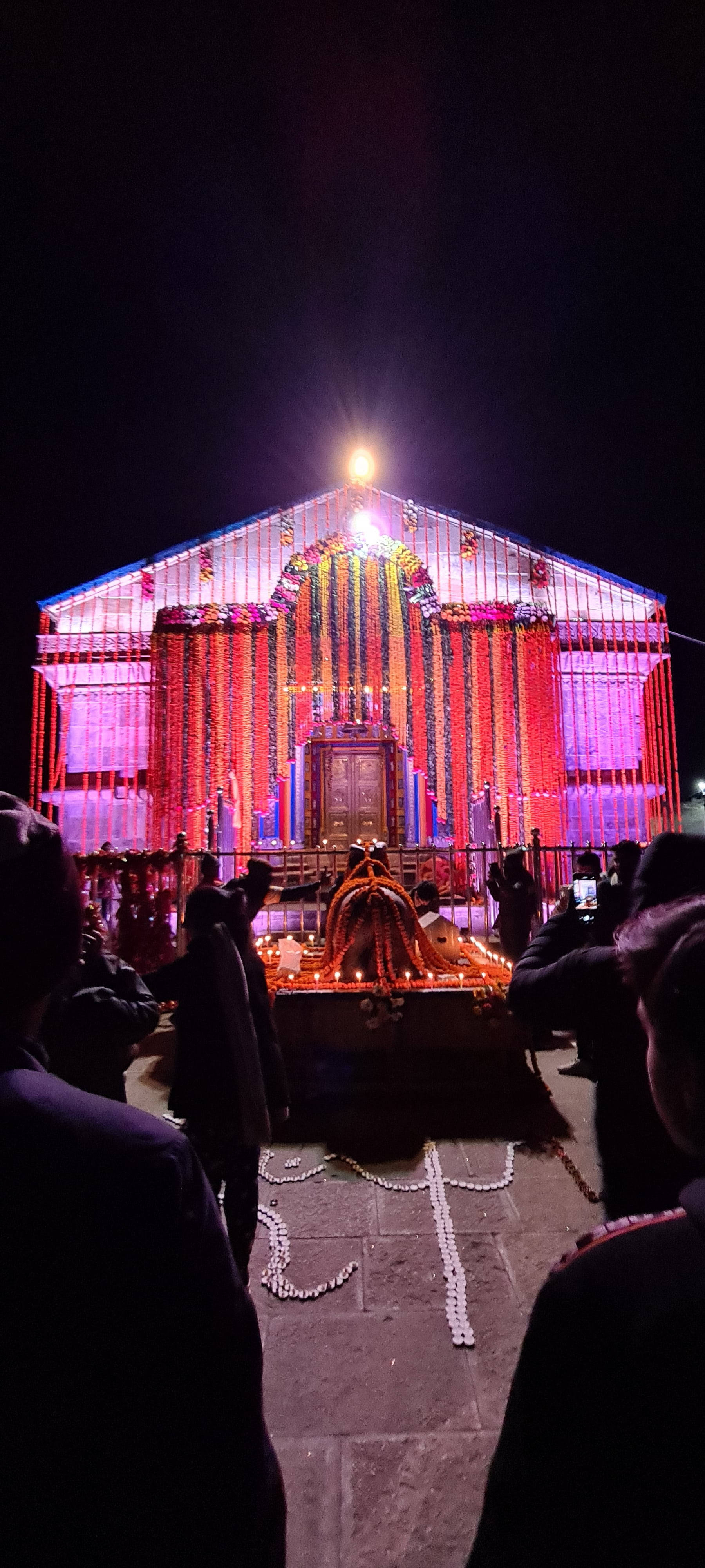 Kedarnath 桌布 壁紙 — 一座宏偉的寺廟在夜晚被鮮豔的粉紅色和紫色燈光照亮，周圍環繞著信徒和儀式供品。