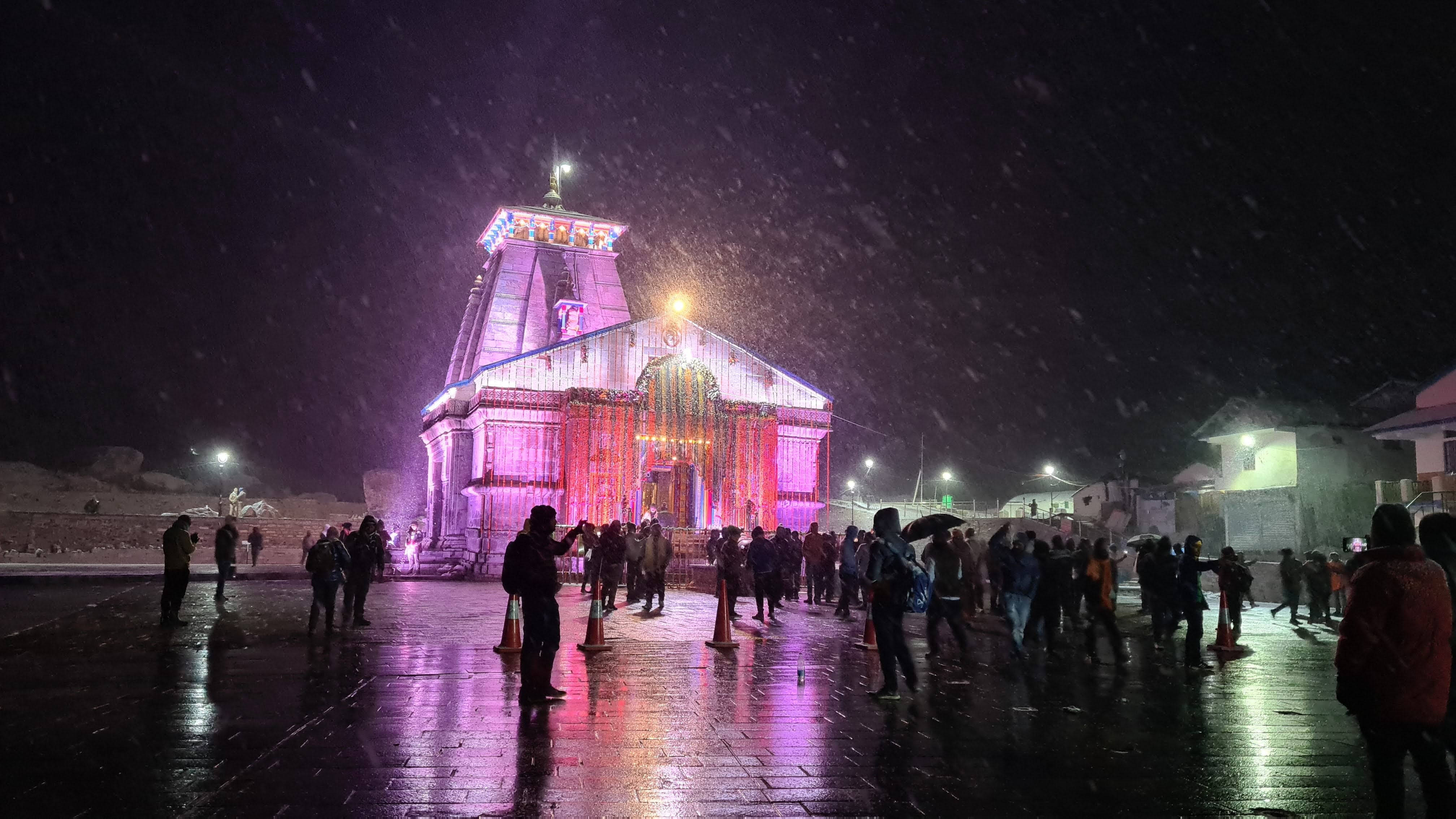 Kedarnath 桌布 壁紙 — 一座宏偉的寺廟在粉紅色和紫色的燈光下閃耀，矗立在飄落的雪中，夜晚濕漉漉的廣場上聚集著剪影般的人群。