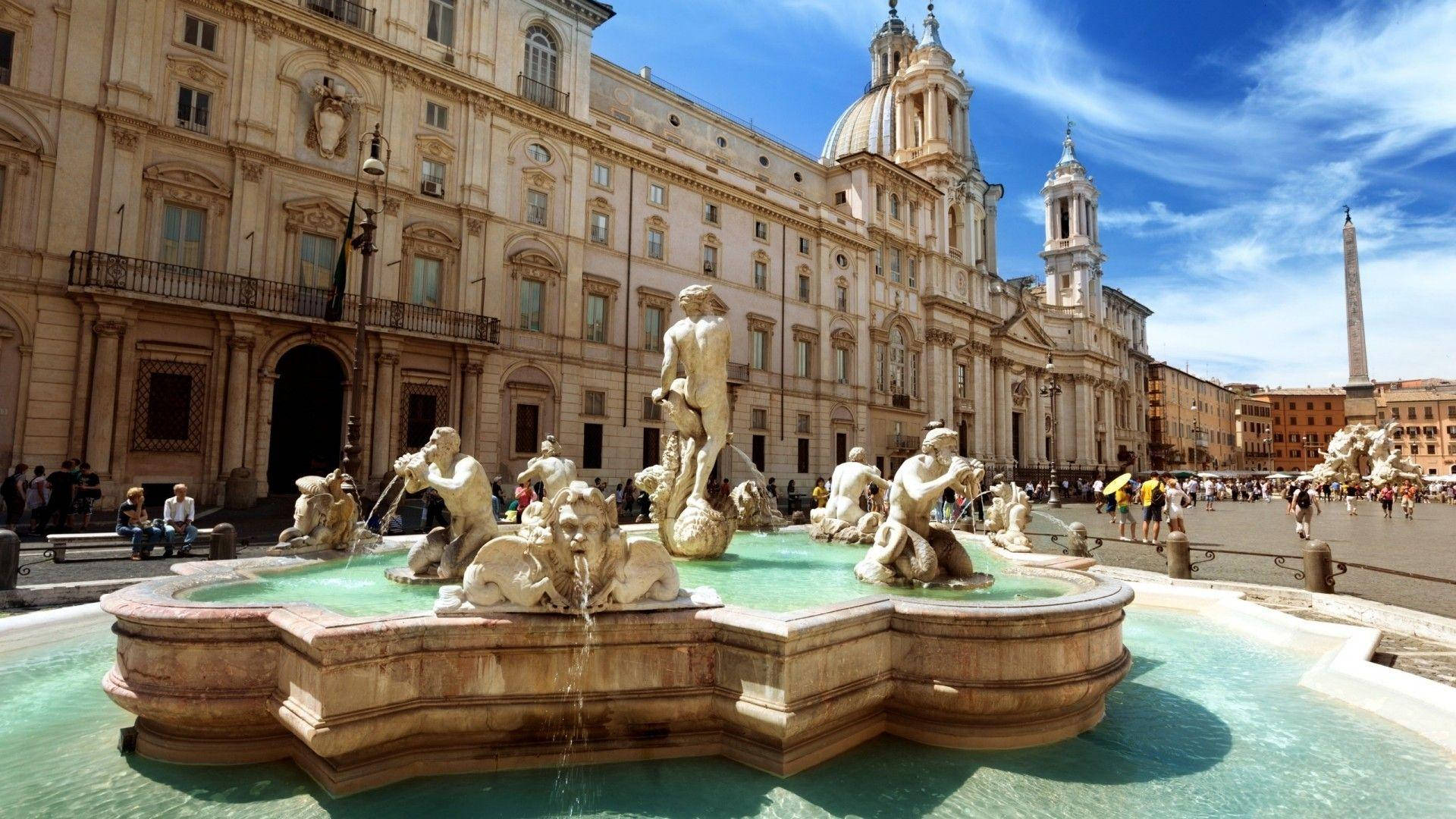 Rome 桌布 壁紙 — 一座宏偉的巴洛克式噴泉，帶有大理石雕像和流動的水，位於陽光明媚的羅馬廣場，兩旁是歷史建築和遠處的方尖碑。