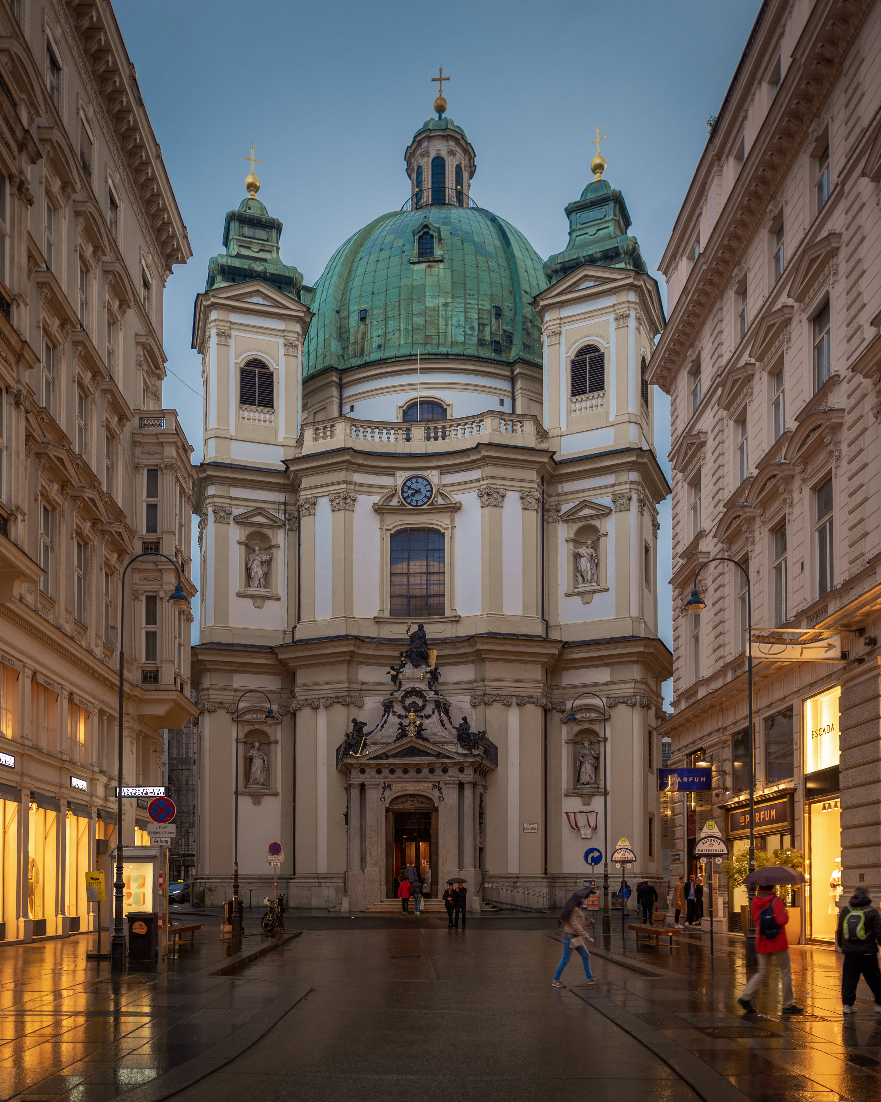 Austria 桌布 壁紙 — 一座宏偉的巴洛克式教堂，帶有綠色銅製圓頂，矗立在潮濕的城市街道盡頭，行人於晚間燈光下走過。