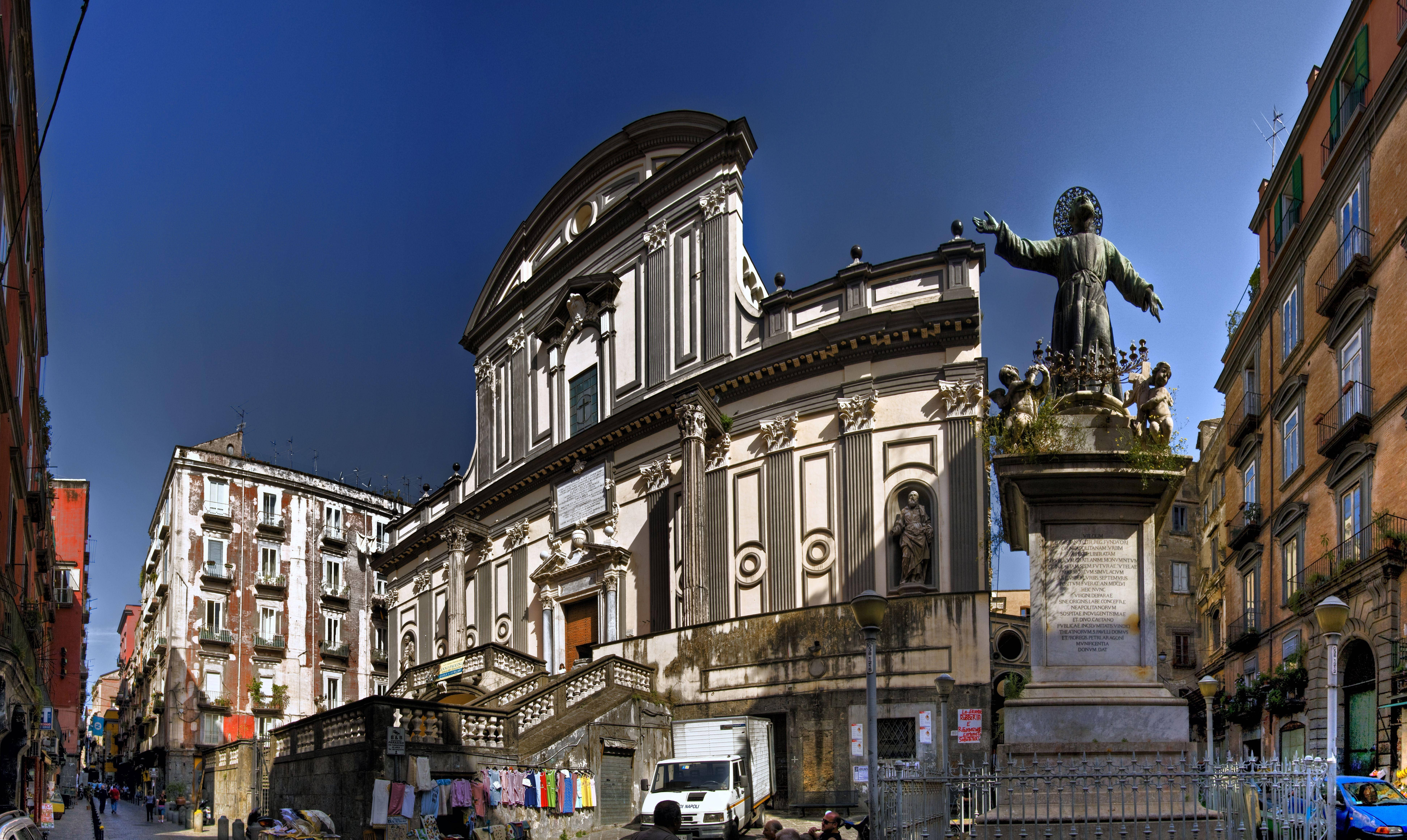 Naples 桌布 壁紙 — 一座宏偉的巴洛克式教堂，擁有華麗的外牆和雕像，矗立在陽光普照的狹窄歐洲街道上，兩旁是色彩繽紛的建築。