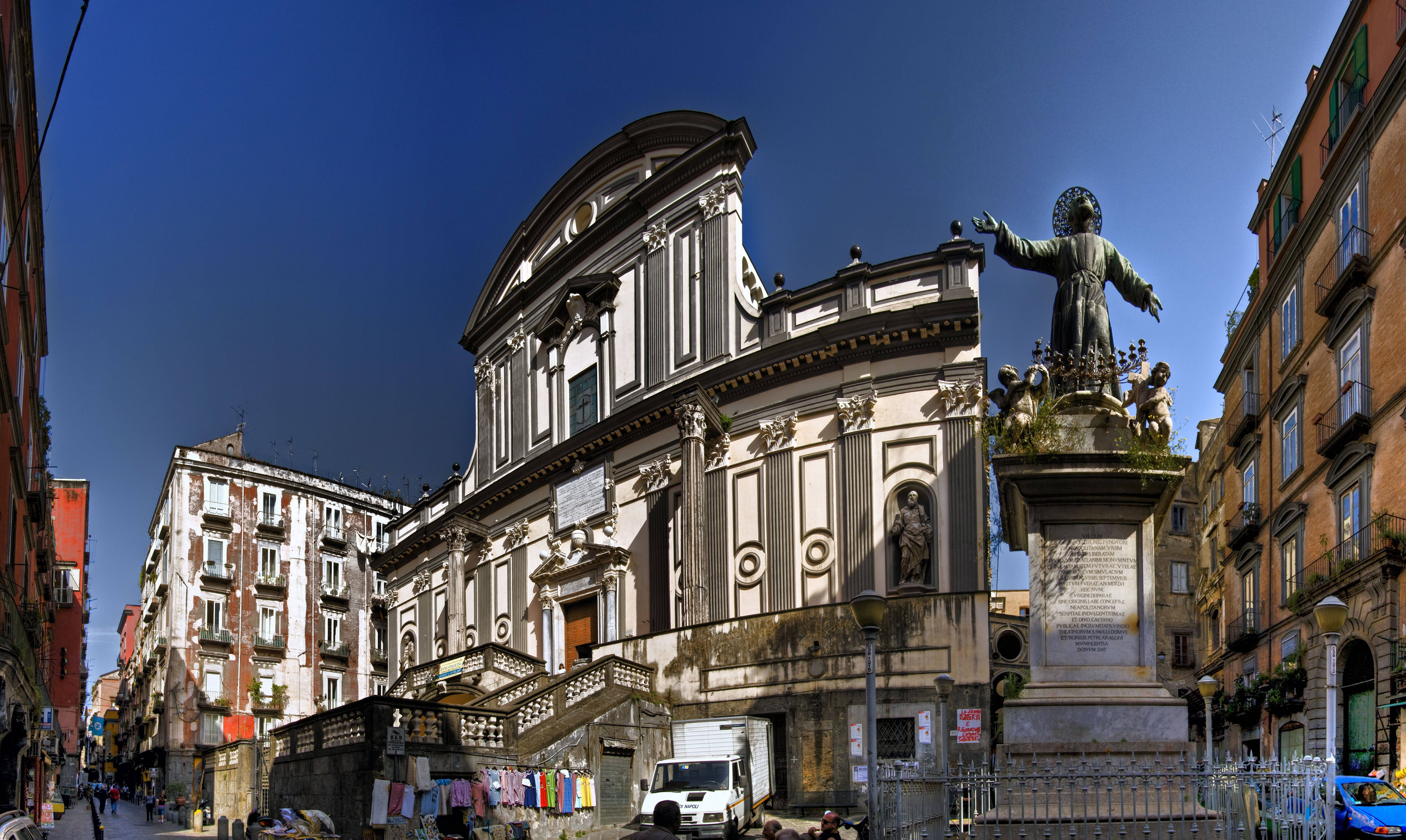 Naples 桌布 壁紙 — 一座宏偉的巴洛克式教堂，擁有華麗的外牆和雕像，矗立在陽光明媚的歐洲街道上，兩旁是色彩繽紛的建築。