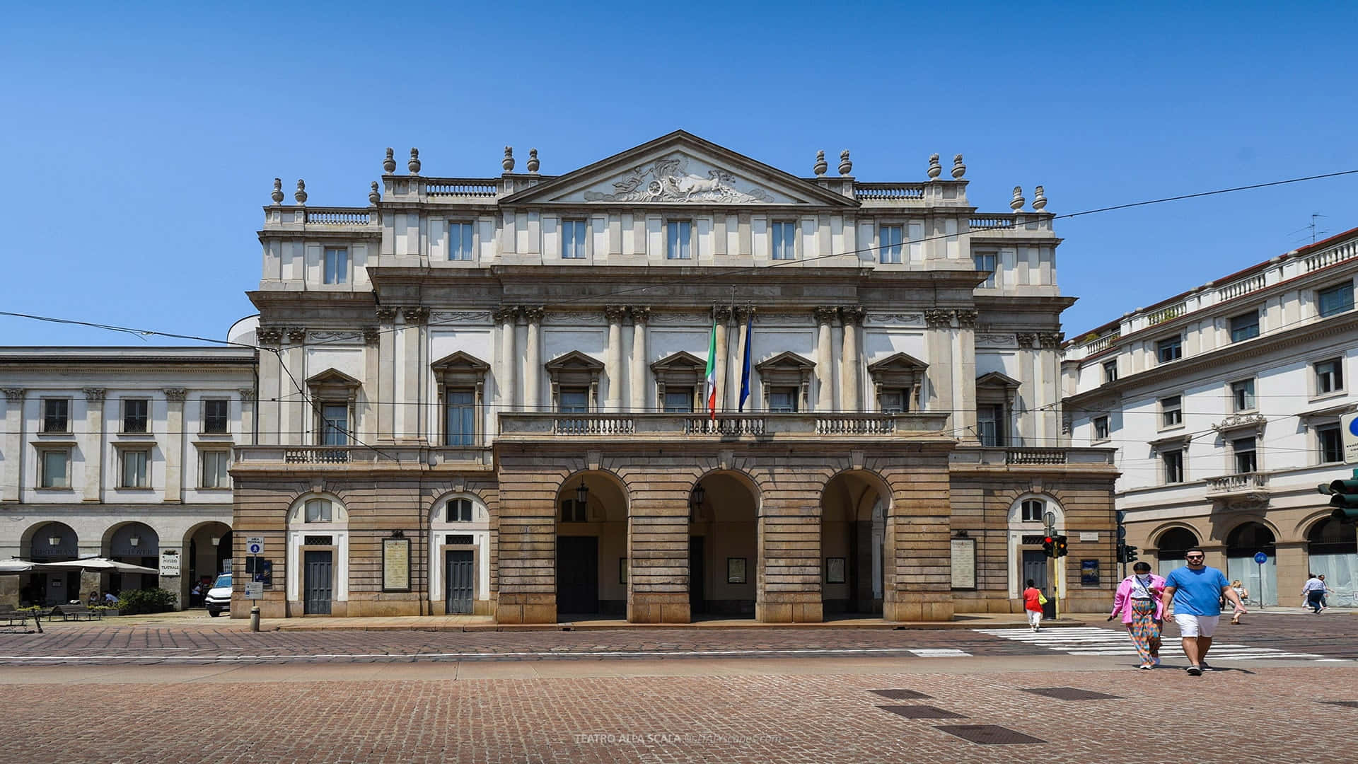 Milan 桌布 壁紙 — 一座宏偉的新古典主義劇院，擁有拱形入口和華麗的外牆，矗立在清澈的藍天下，行人漫步在陽光普照的磚砌廣場上。
