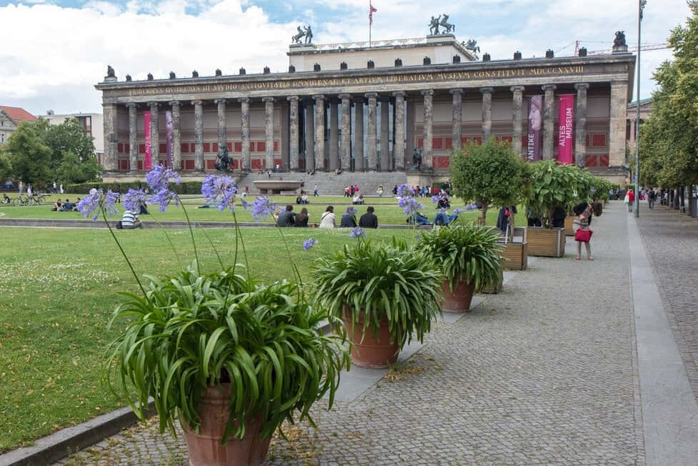 Berlin 桌布 壁紙 — 一座宏偉的新古典主義博物館，擁有柱子和雕像，俯瞰著一個熱鬧的公園，人們在部分多雲的天空下在草地上放鬆。